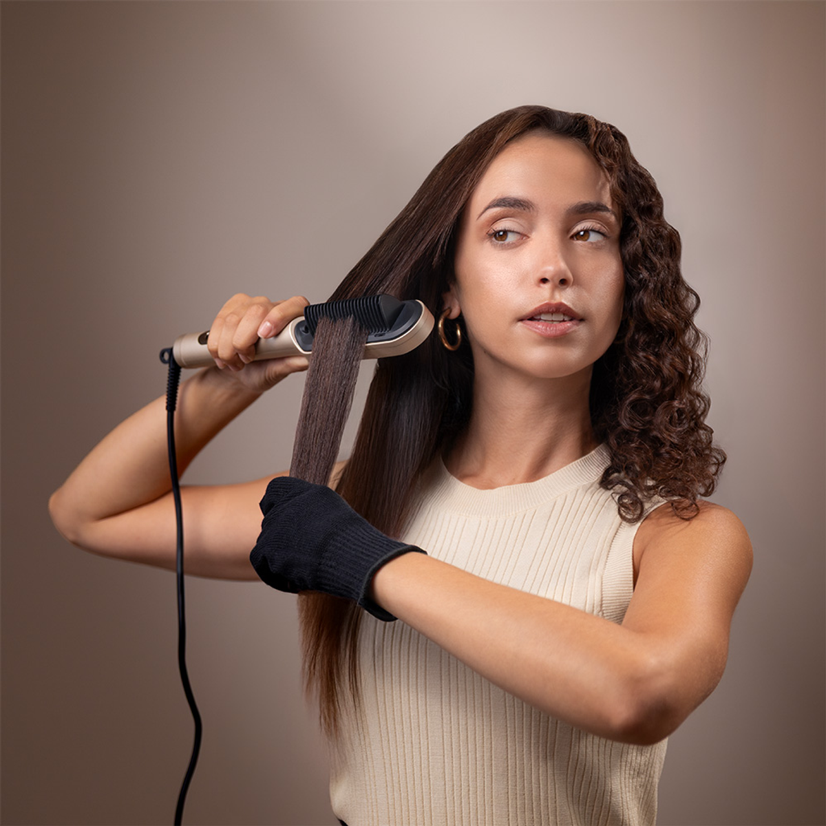 Donna che si liscia i capelli con una piastra. Un lato è liscio, l'altro riccio. Indossa un top color crema e un guanto resistente al calore.