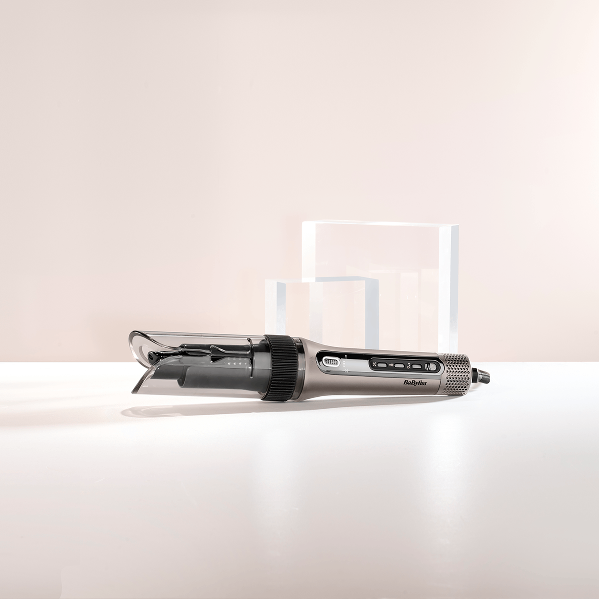 Szara lokówka BaByliss na białej powierzchni. Dwa przezroczyste bloki w tle, na jasnoróżowym tle.
