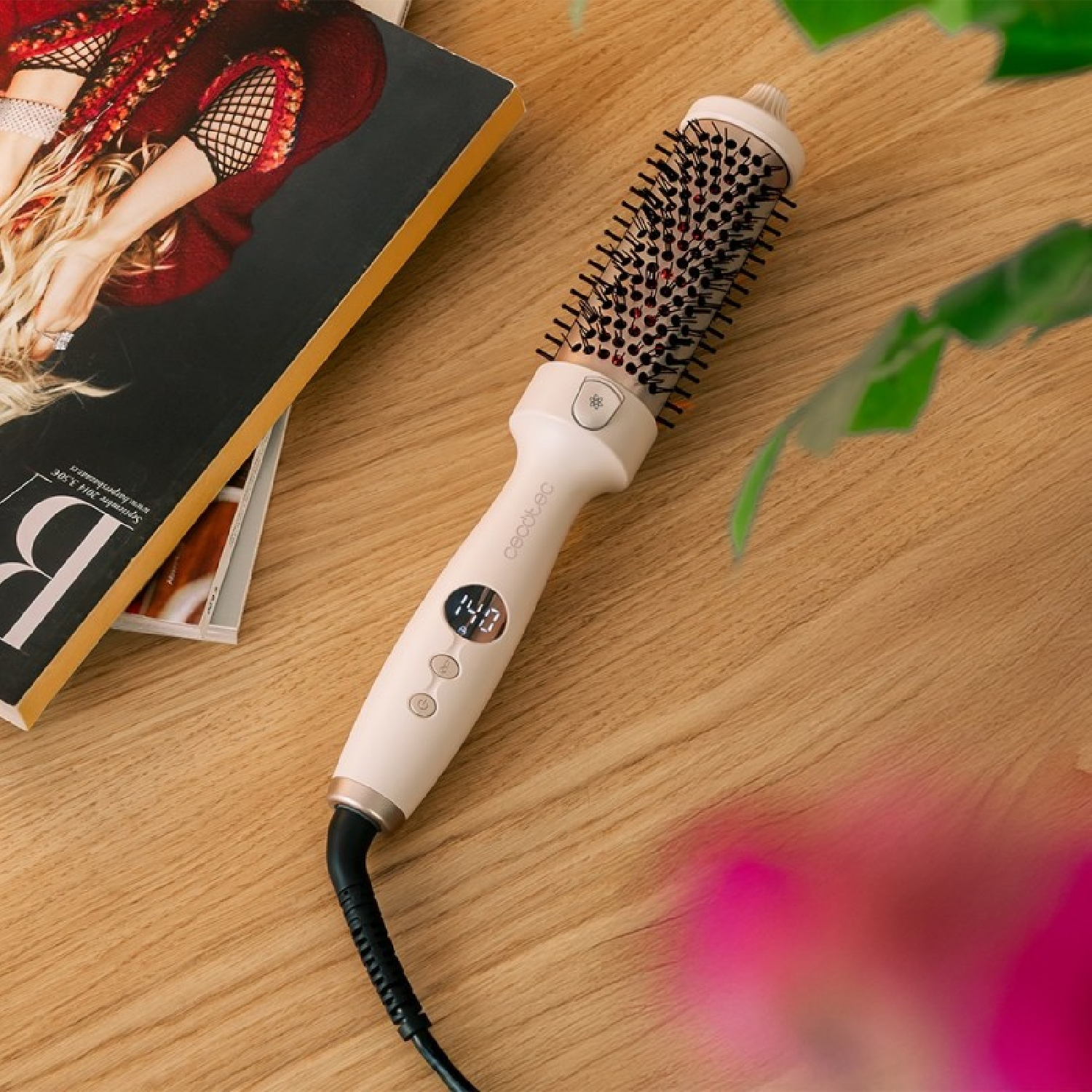 Una spazzola per capelli bianca e nera con display digitale, appoggiata su una superficie di legno accanto a una rivista e foglie verdi.