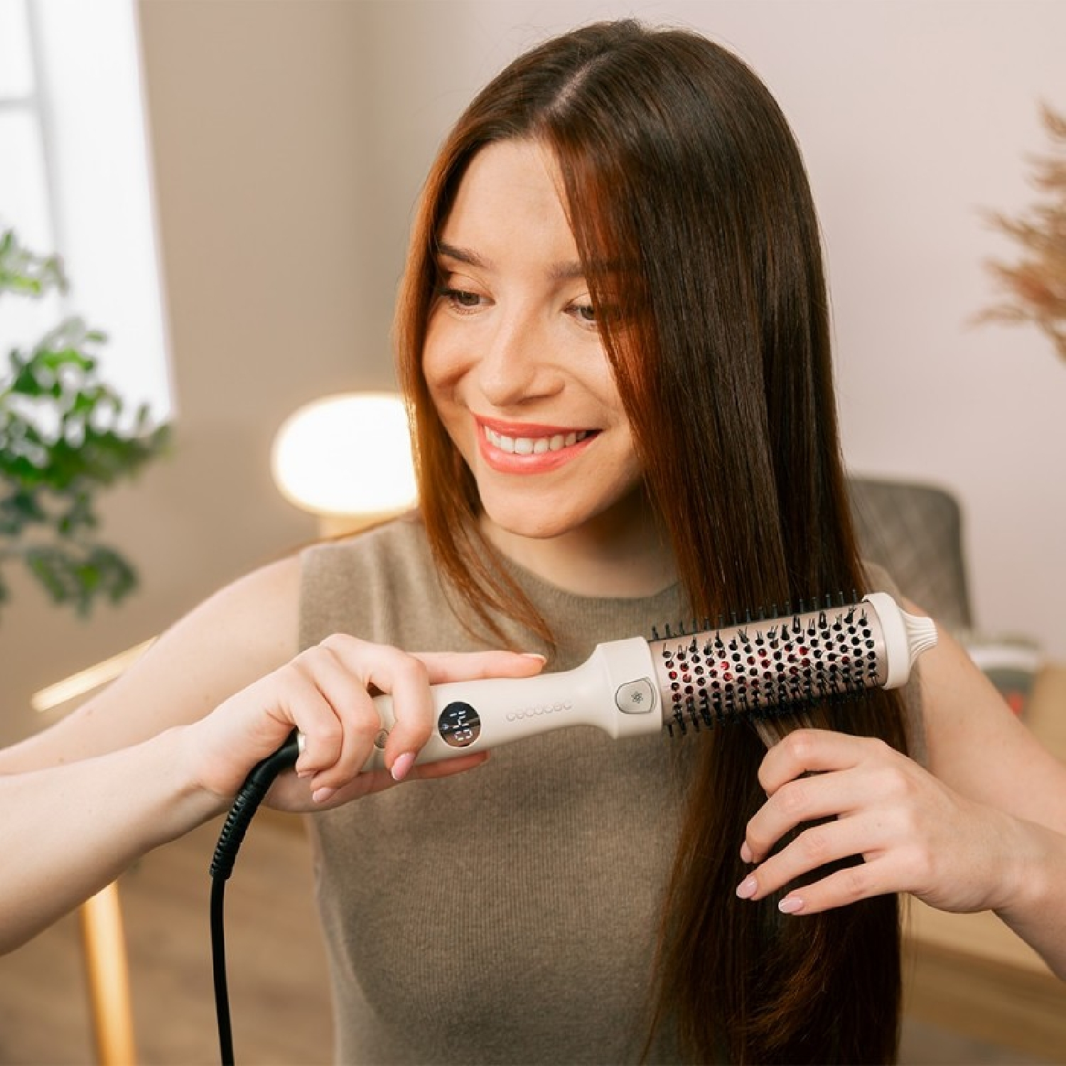 Una donna con lunghi capelli castani sorride mentre usa una spazzola per capelli. È in una stanza con piante sullo sfondo.