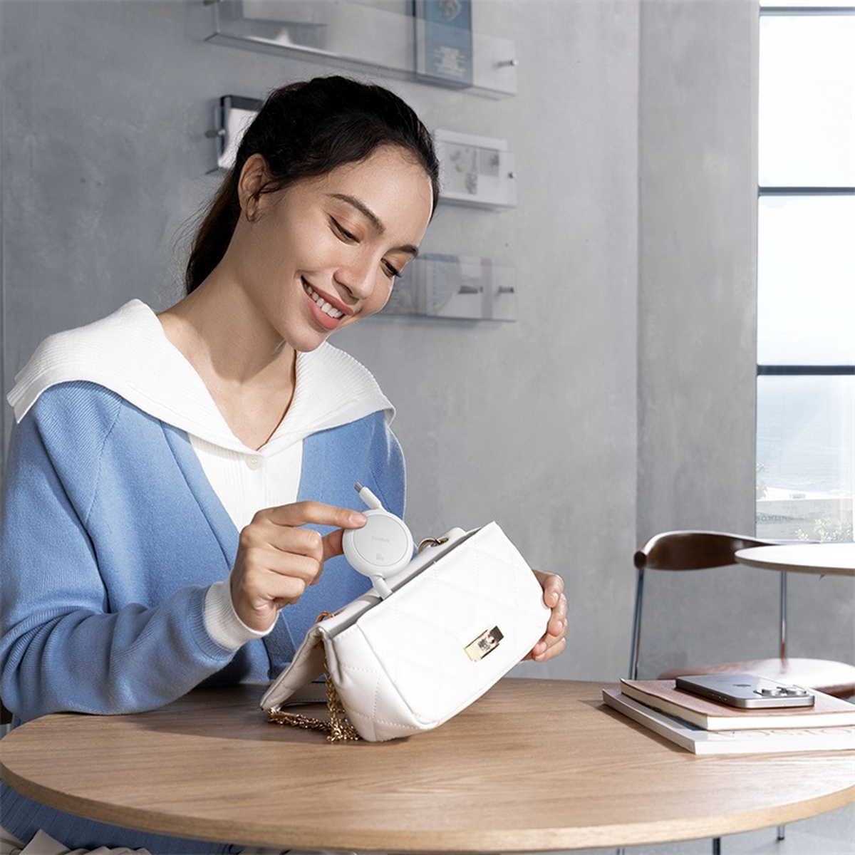 Eine lächelnde Frau hält ein weißes Gerät und legt es in eine weiße Handtasche. Ein Holztisch mit Büchern und einem Telefon vor ihr.