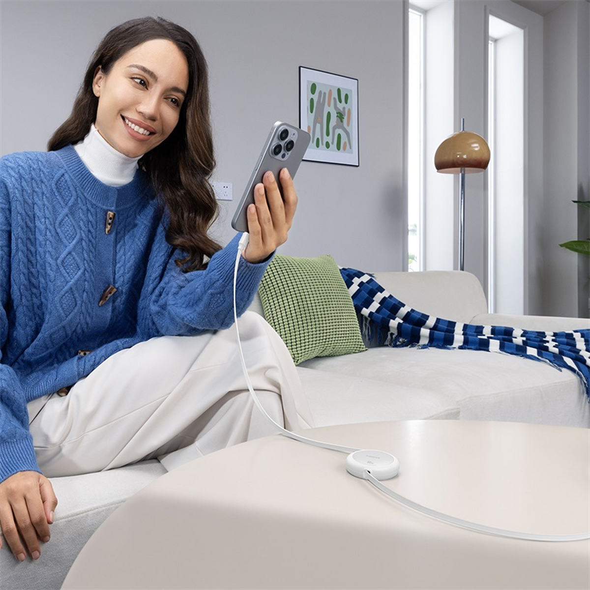 Frau hält ein Telefon, das mit einem weißen Ladegerät verbunden ist. Sie trägt einen blauen Pullover und sitzt auf einem weißen Sofa. Ein Gemälde hängt an der Wand.