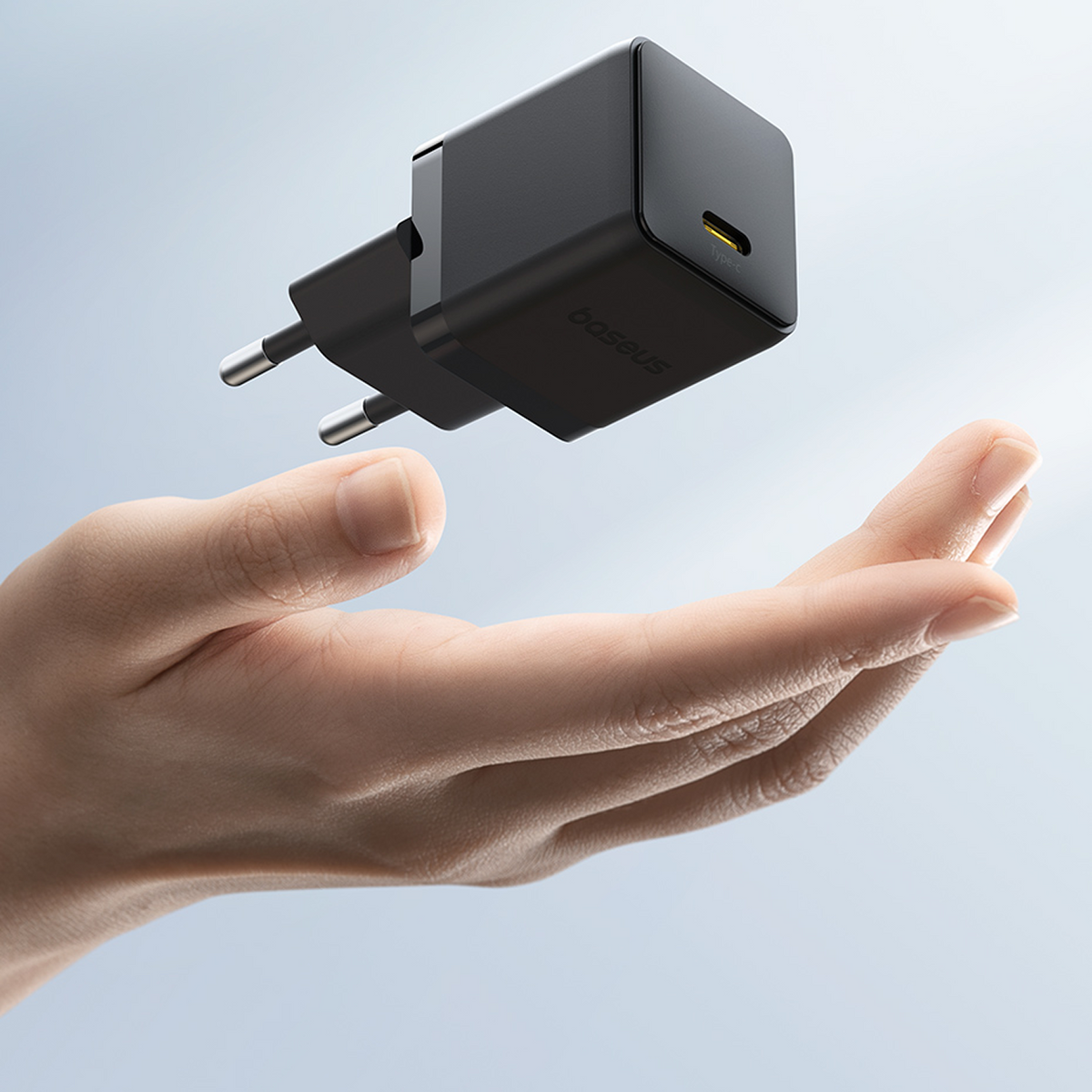 Un caricabatterie Baseus nero fluttua sopra una mano aperta. Ha due spine e una porta USB-C. Lo sfondo è sfocato.
