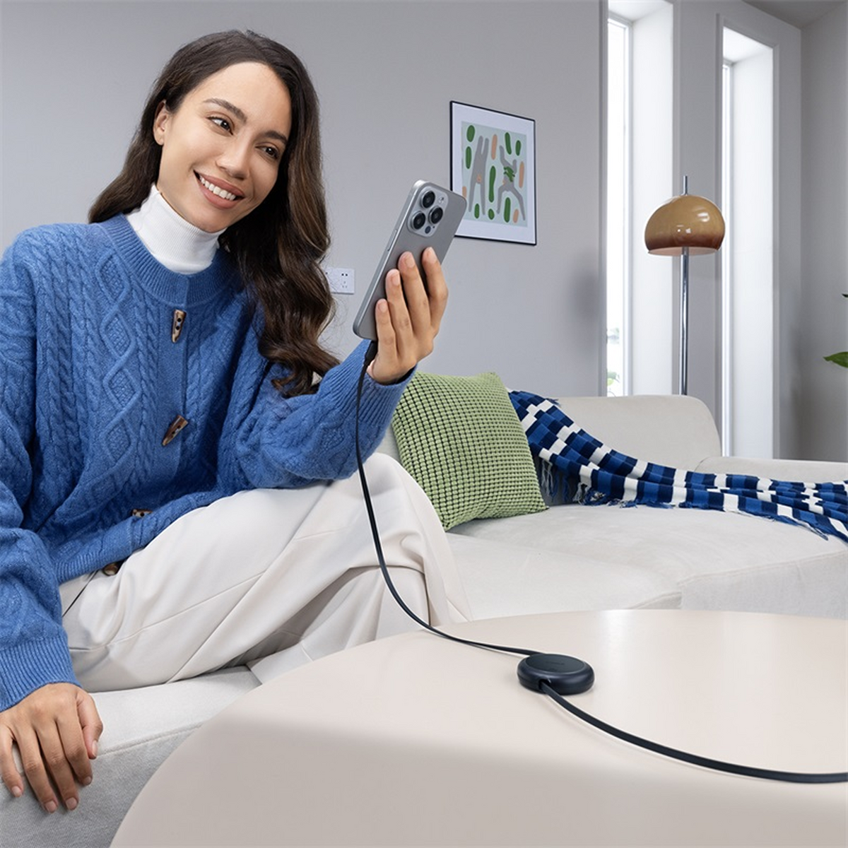 Woman in blue sweater holding phone, connected to a charger on a table. Couch and lamp in background.