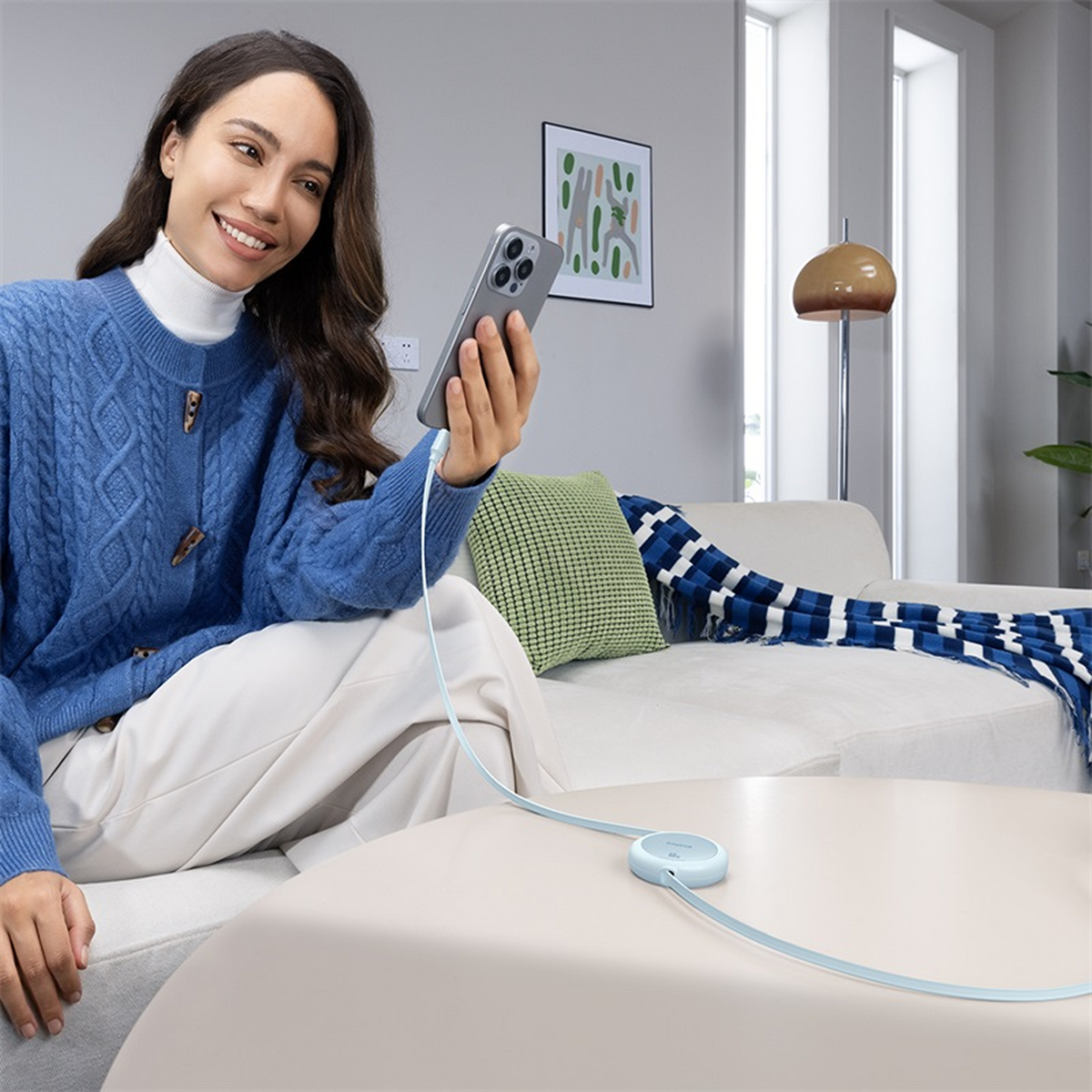 Eine Frau lächelt, während sie ein Telefon hält, das mit einem Kabel und einem Ladehub auf einem Tisch verbunden ist. Im Hintergrund sind eine Couch und eine Lampe zu sehen.
