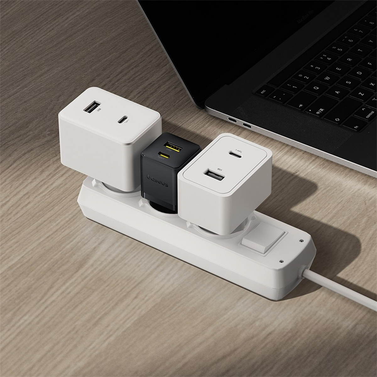 Une multiprise avec trois chargeurs USB branchés. Un ordinateur portable est en arrière-plan. Les chargeurs sont blancs et noirs.
