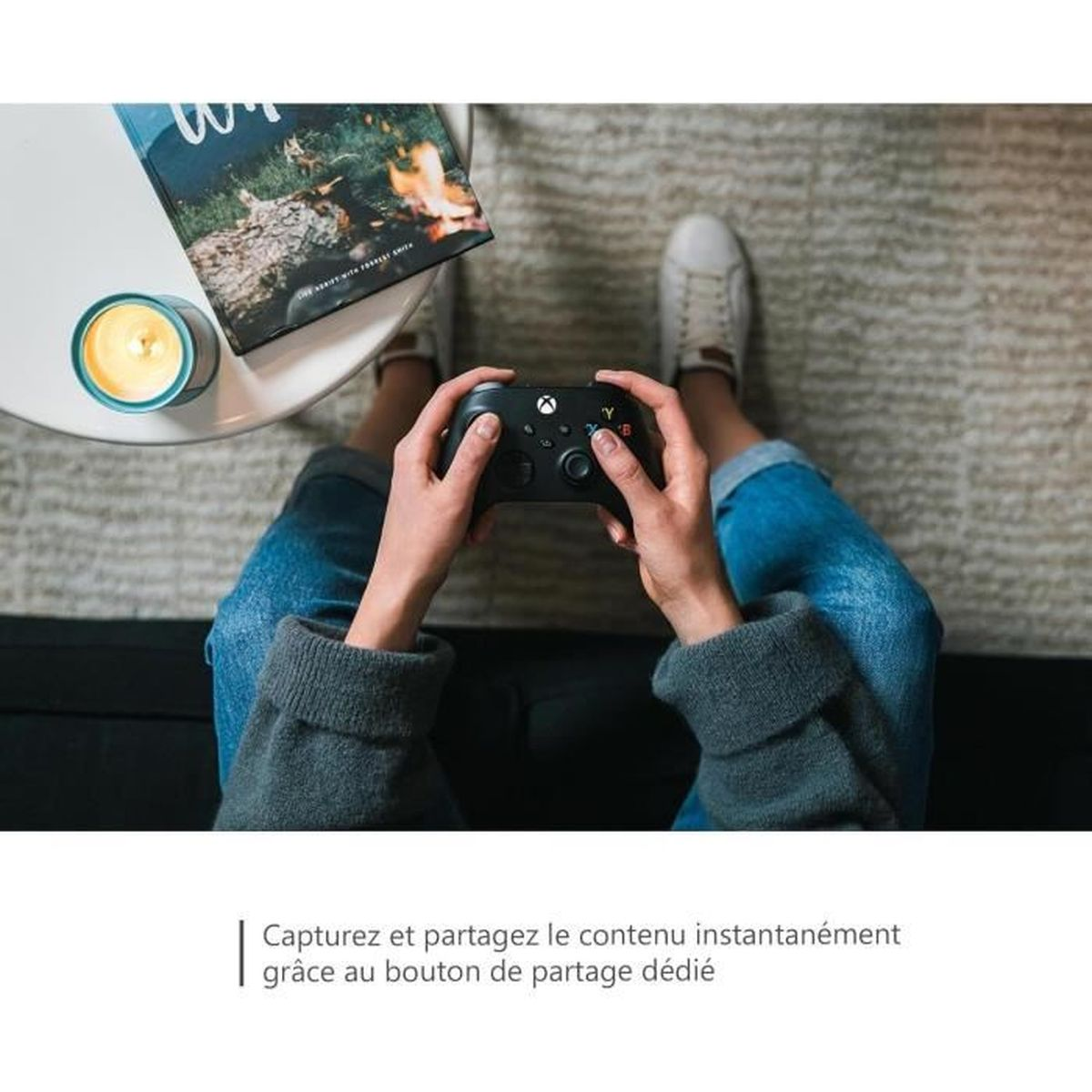 Persona che tiene in mano un controller Xbox nero. Un tavolo bianco con candela e libro è accanto alle gambe. Jeans e scarpe bianche.