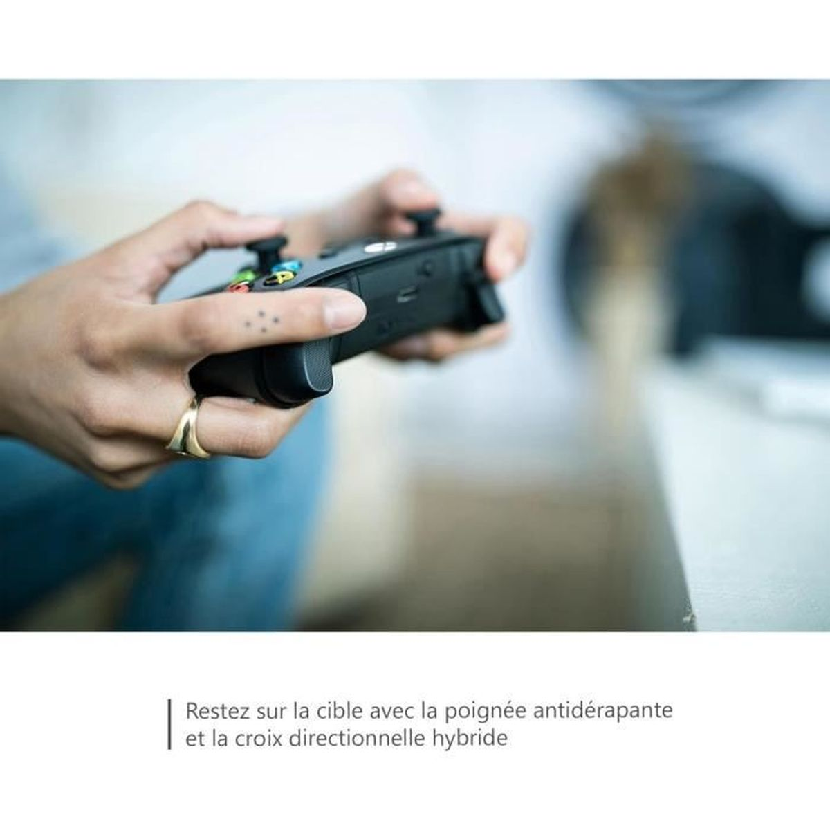 Una persona tiene in mano un controller di gioco nero. Le sue mani sono visibili, con un anello d'oro e un tatuaggio. Lo sfondo è sfocato.