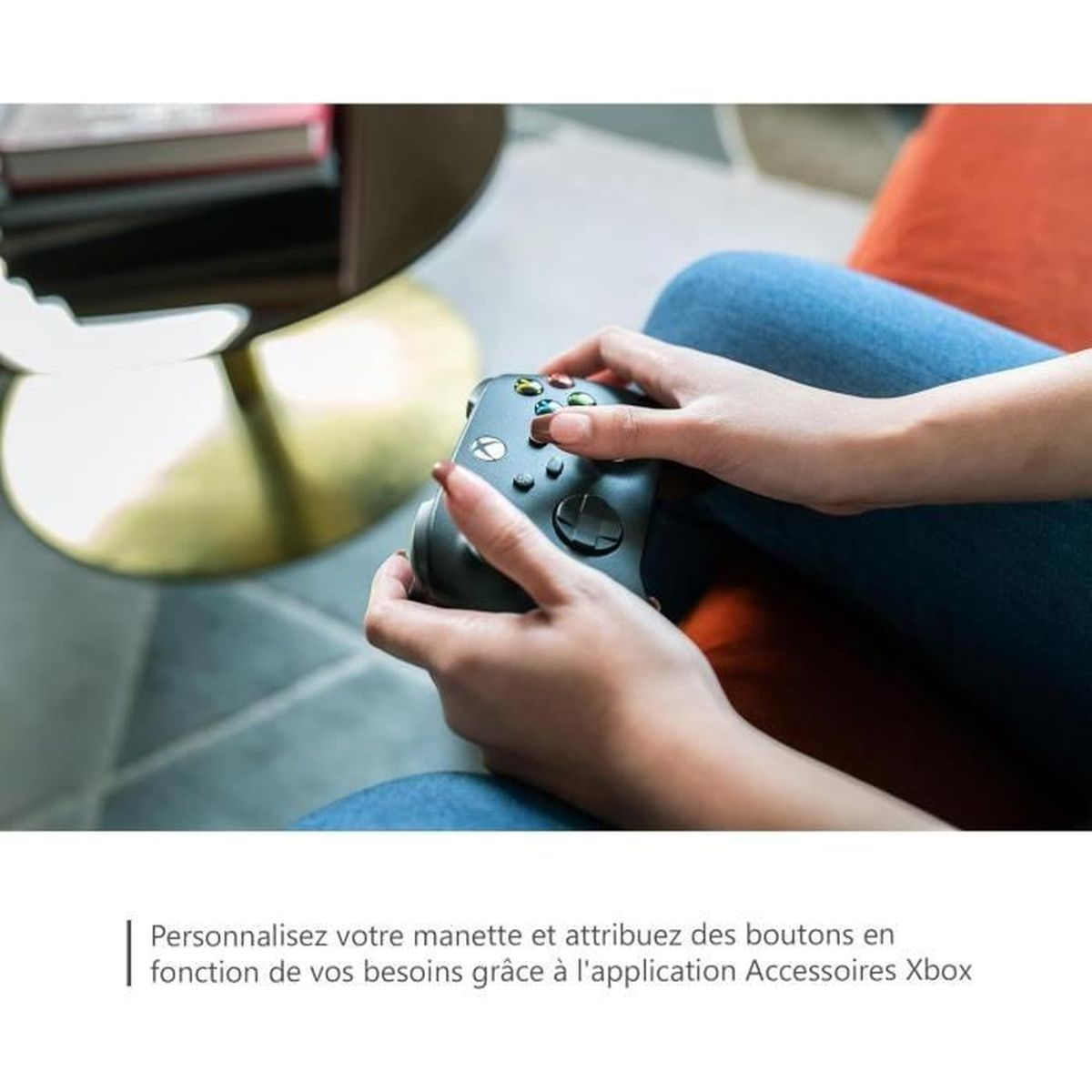 Mani che tengono un controller Xbox nero. I pulsanti sono visibili. Jeans e sedia sullo sfondo. Presenti anche un tavolo e libri.