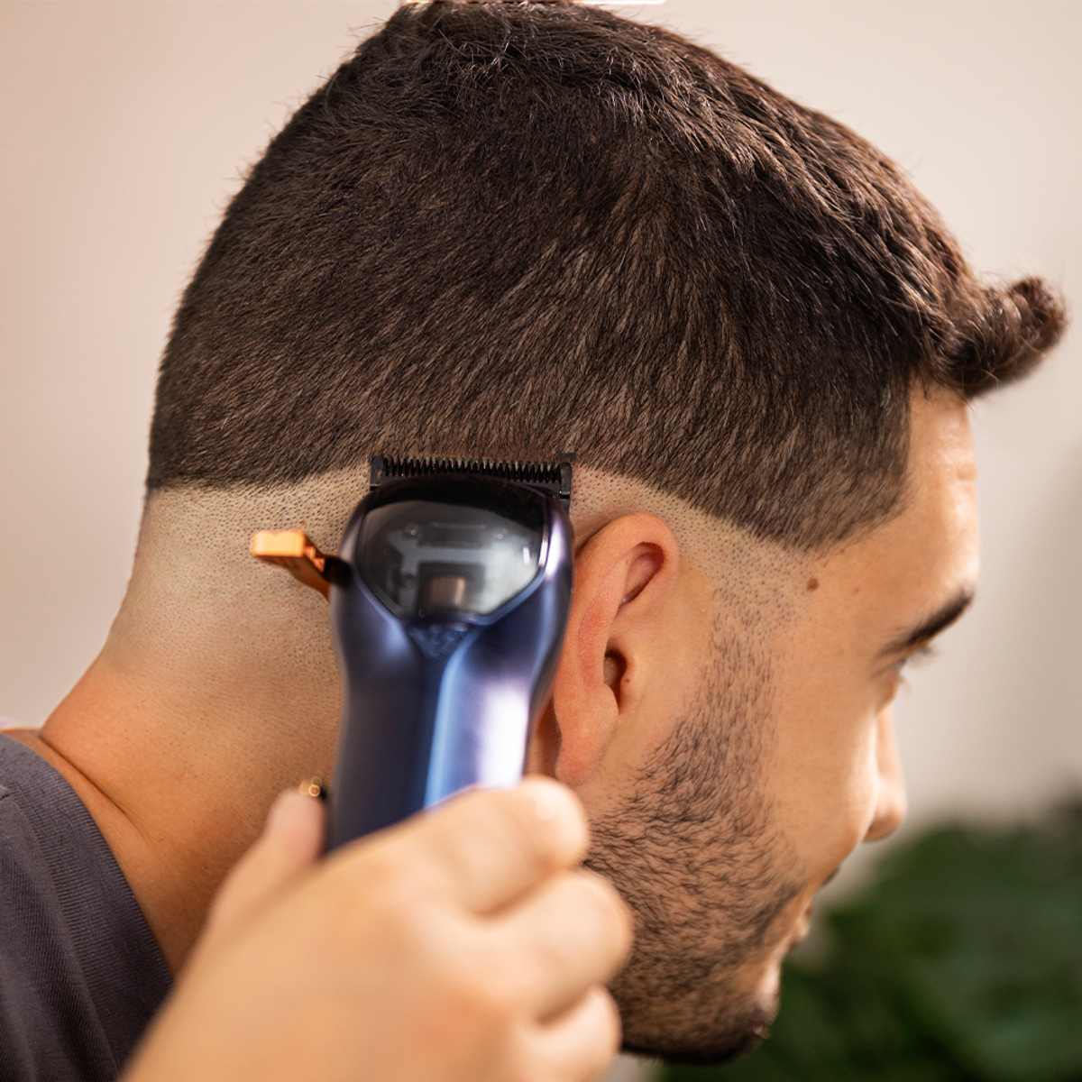 Un uomo si fa tagliare i capelli con un tagliacapelli. Il tagliacapelli è blu scuro e oro. L'uomo ha i capelli scuri e la barba. Lo sfondo è sfocato.