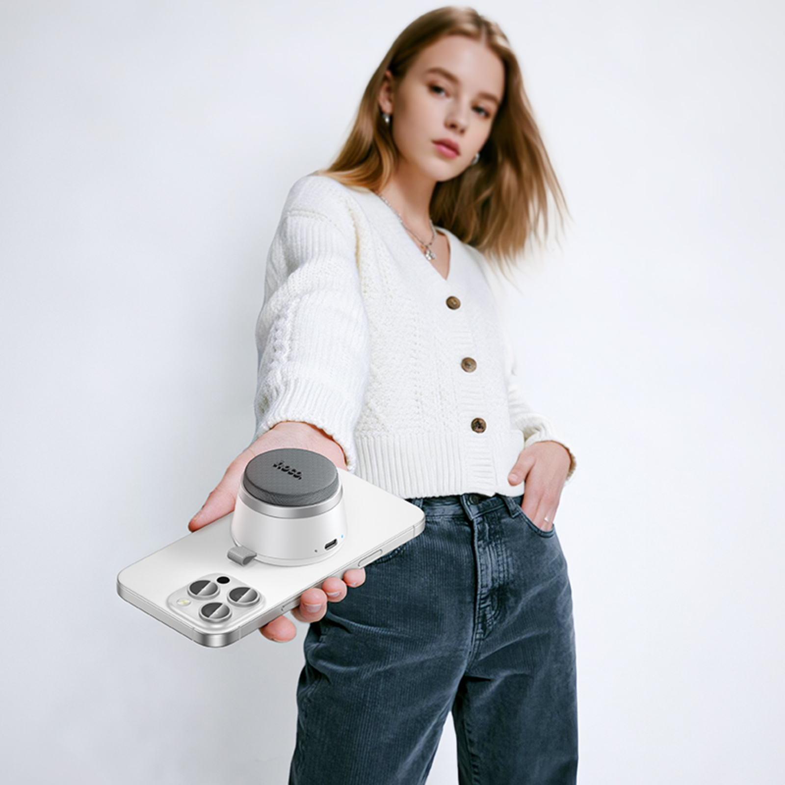 Eine Frau hält ein Telefon mit einem Lautsprecher. Sie trägt einen weißen Pullover und dunkle Jeans vor weißem Hintergrund.