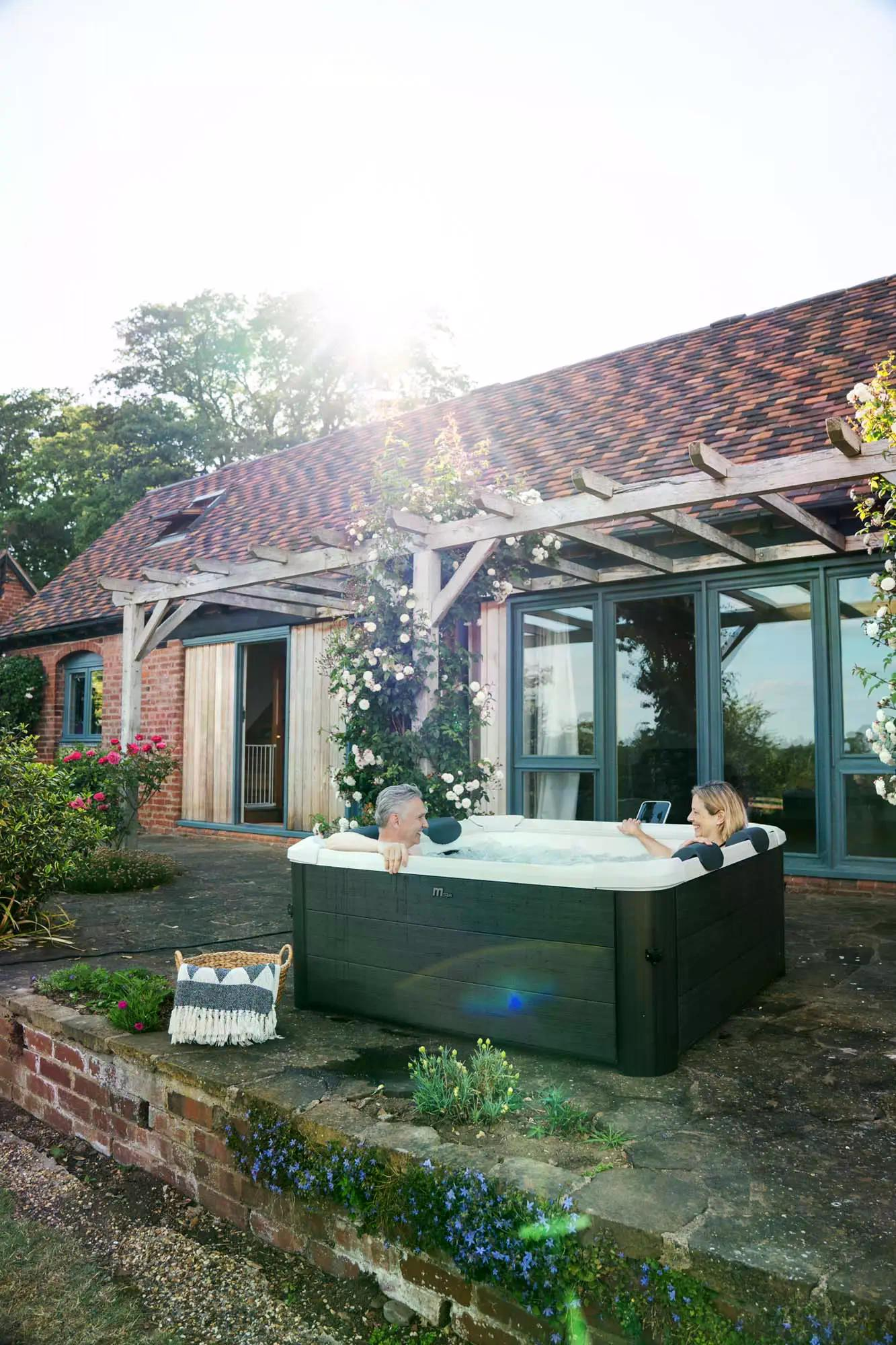 Ein Paar entspannt sich im Freien in einem Whirlpool neben einem Backsteingebäude. Blumen und Grün umgeben die Szene unter sonnigem Himmel.