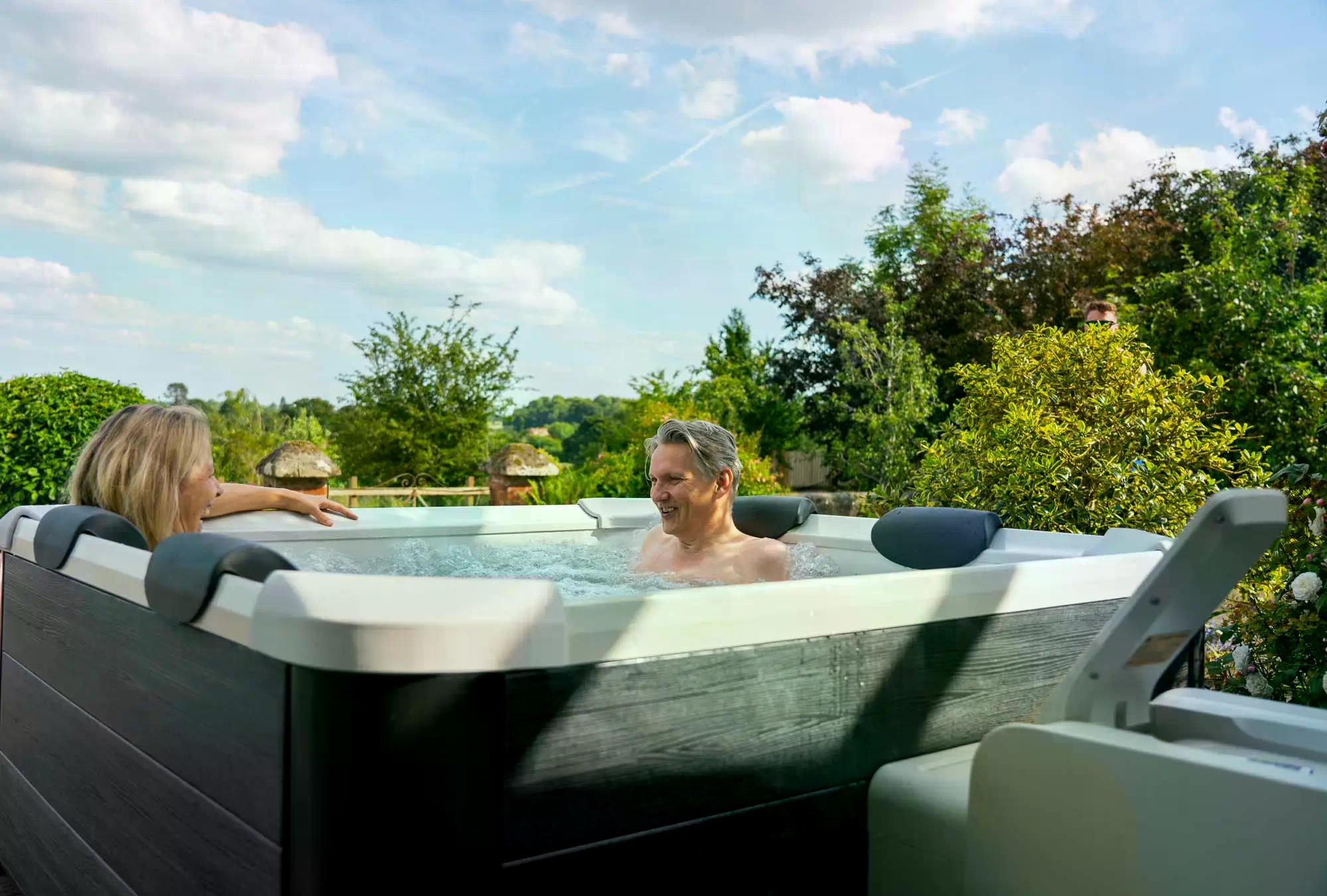 Zwei Personen in einem Whirlpool im Freien. Mann lächelt, Frau schaut. Grüne Bäume und blauer Himmel mit Wolken im Hintergrund.