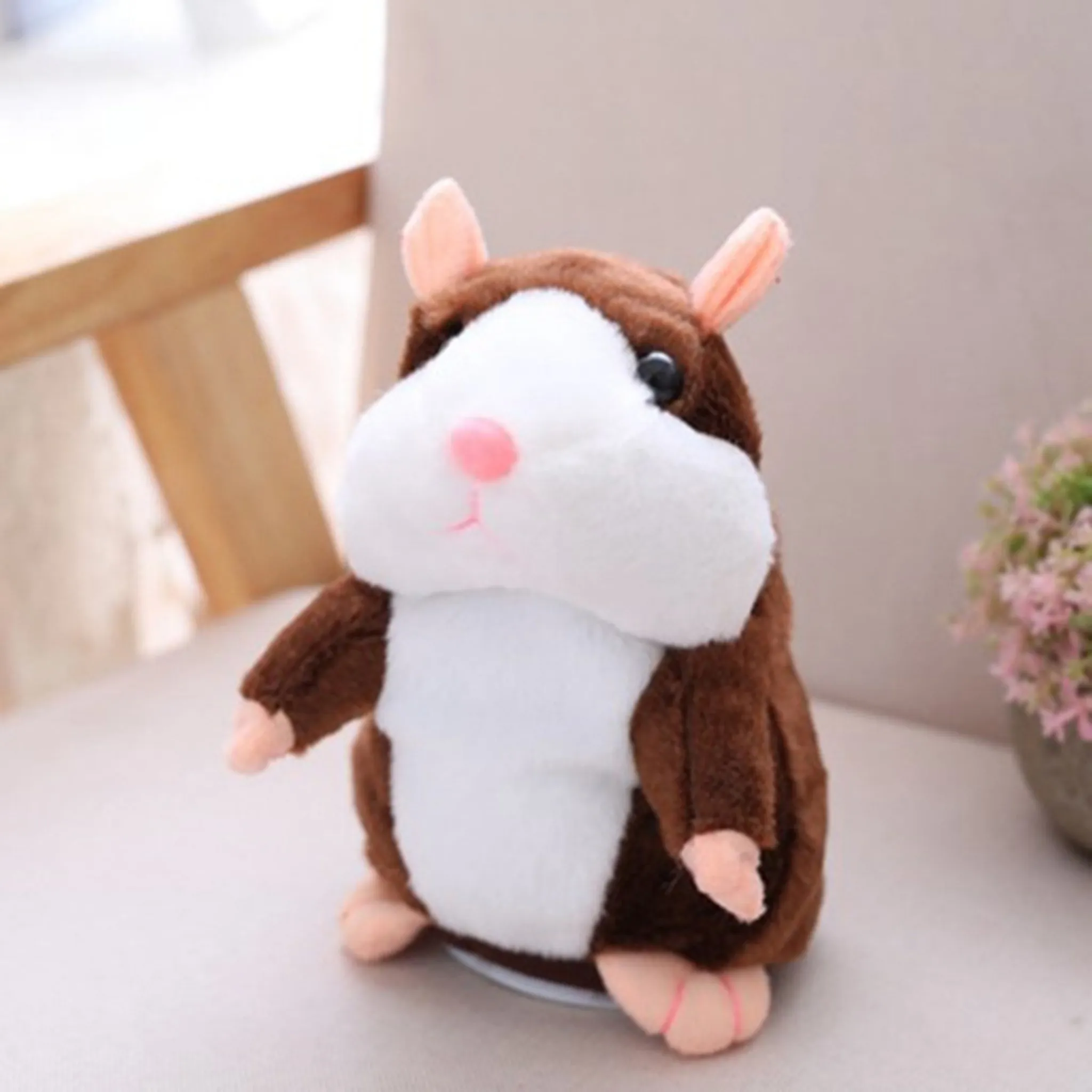 Ein braunes und weißes Hamster-Plüschtier mit rosa Nase und Ohren sitzt auf einer weißen Oberfläche. Blumen sind im Hintergrund.
