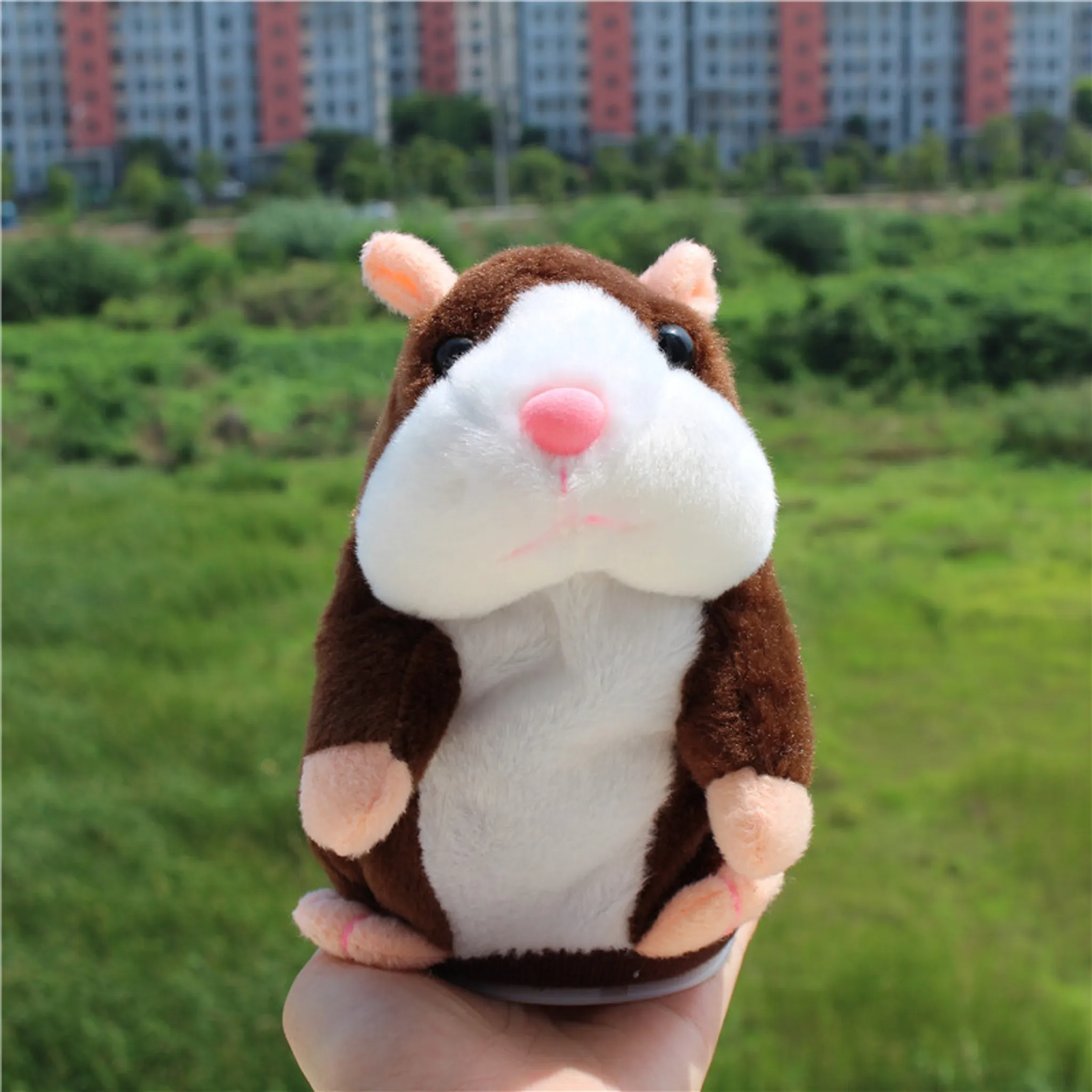 Ein braunes und weißes Hamsterspielzeug mit rosa Nase und Ohren, gehalten in der Hand, mit grünem Hintergrund und Gebäuden.