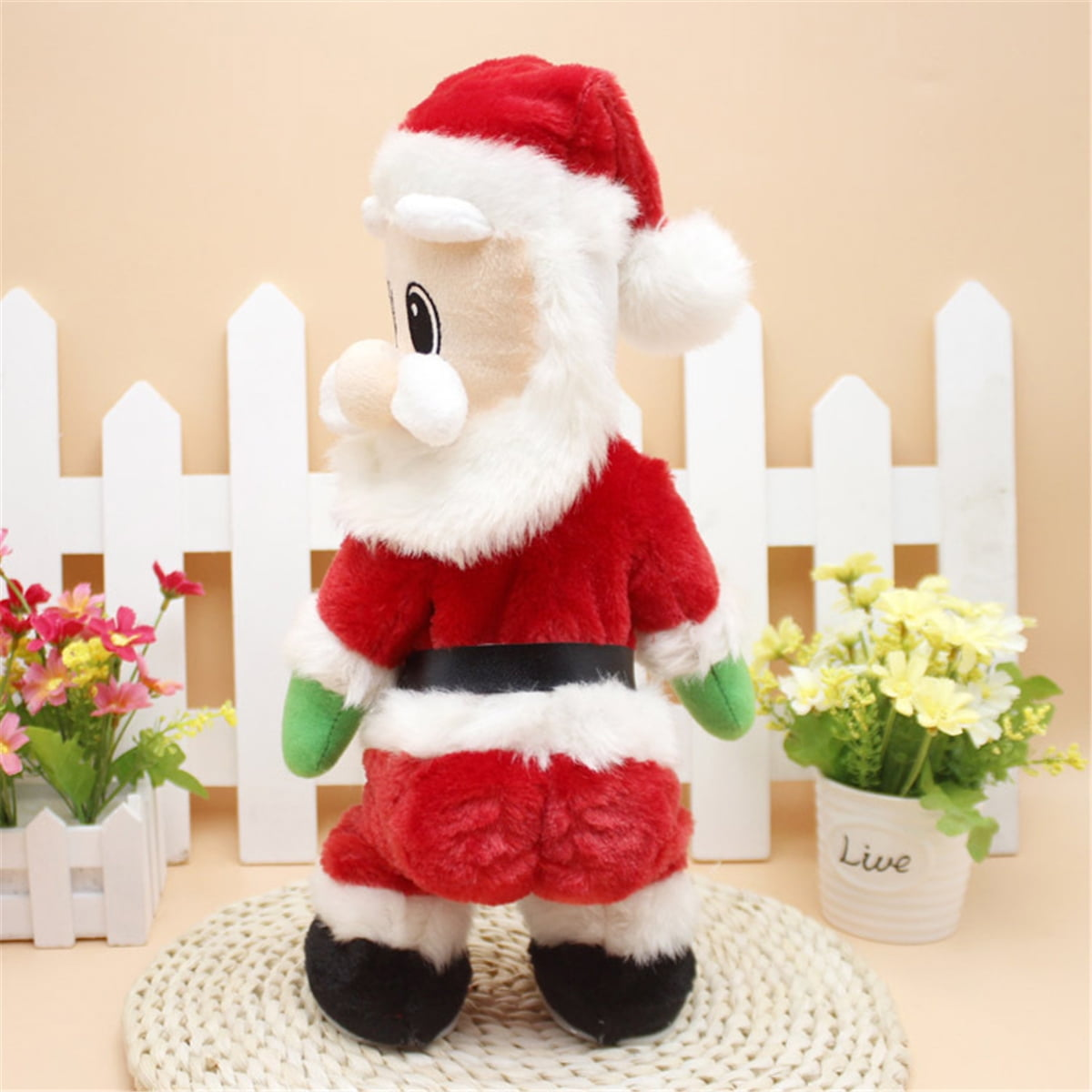 Eine Weihnachtsmann-Puppe mit rotem Mantel, Hut und Hose steht auf einer gewebten Matte. Auf beiden Seiten befinden sich Blumen und ein weißer Zaun.
