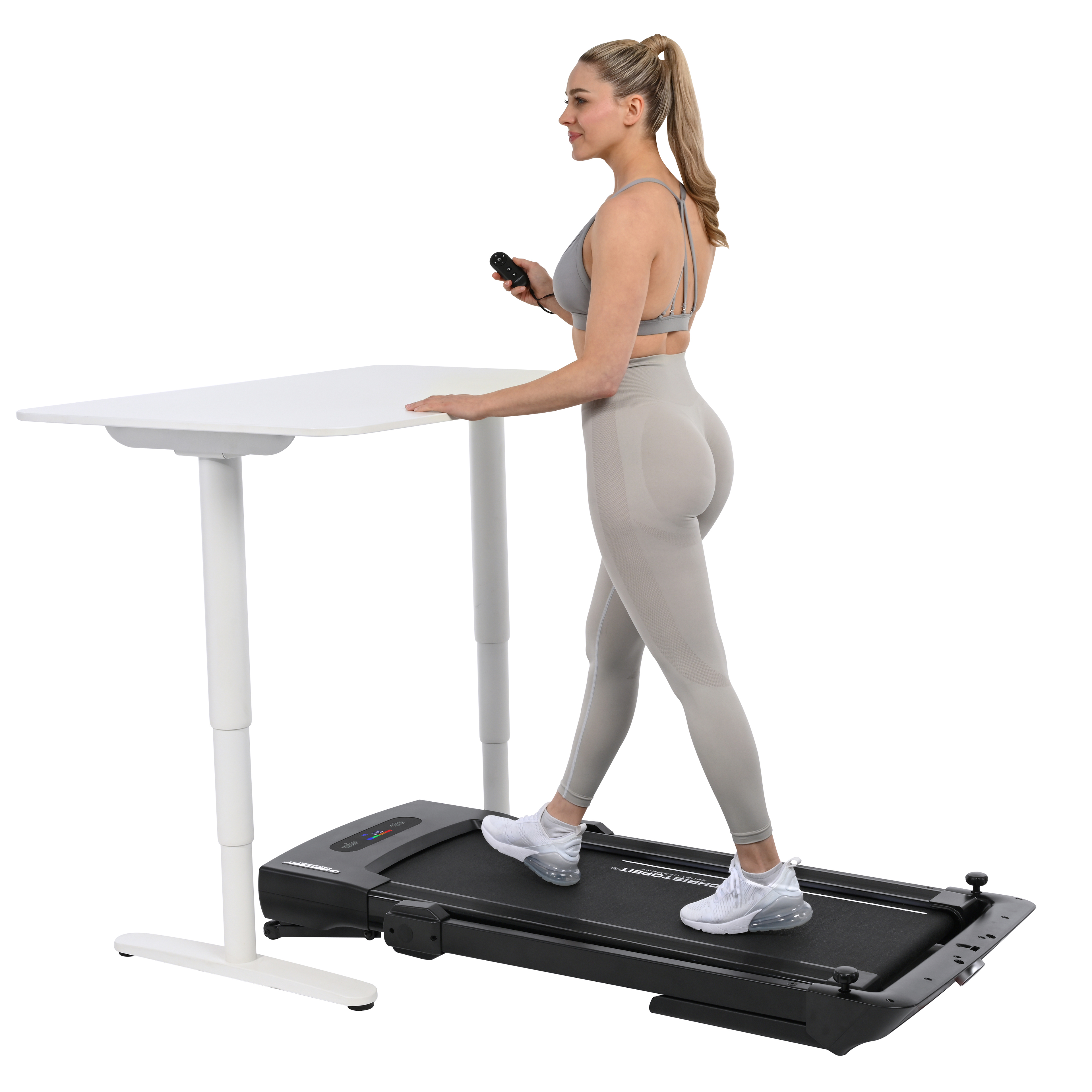 Eine Frau in Sportkleidung geht auf einem Laufband neben einem weißen Schreibtisch und hält eine Fernbedienung.