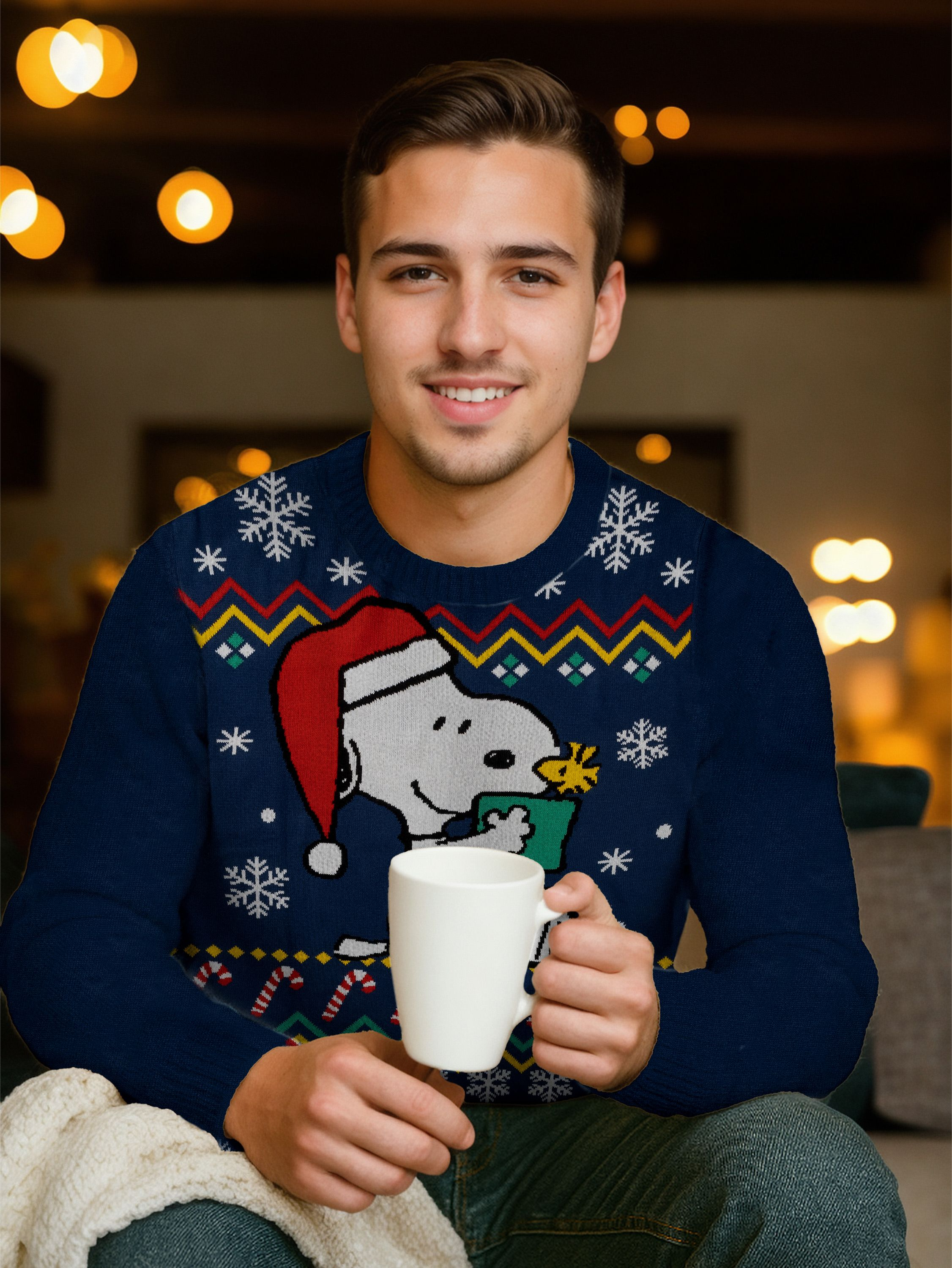 Ein Mann in einem Snoopy-Pullover hält eine weiße Tasse und lächelt. Der Hintergrund hat Bokeh-Lichter.