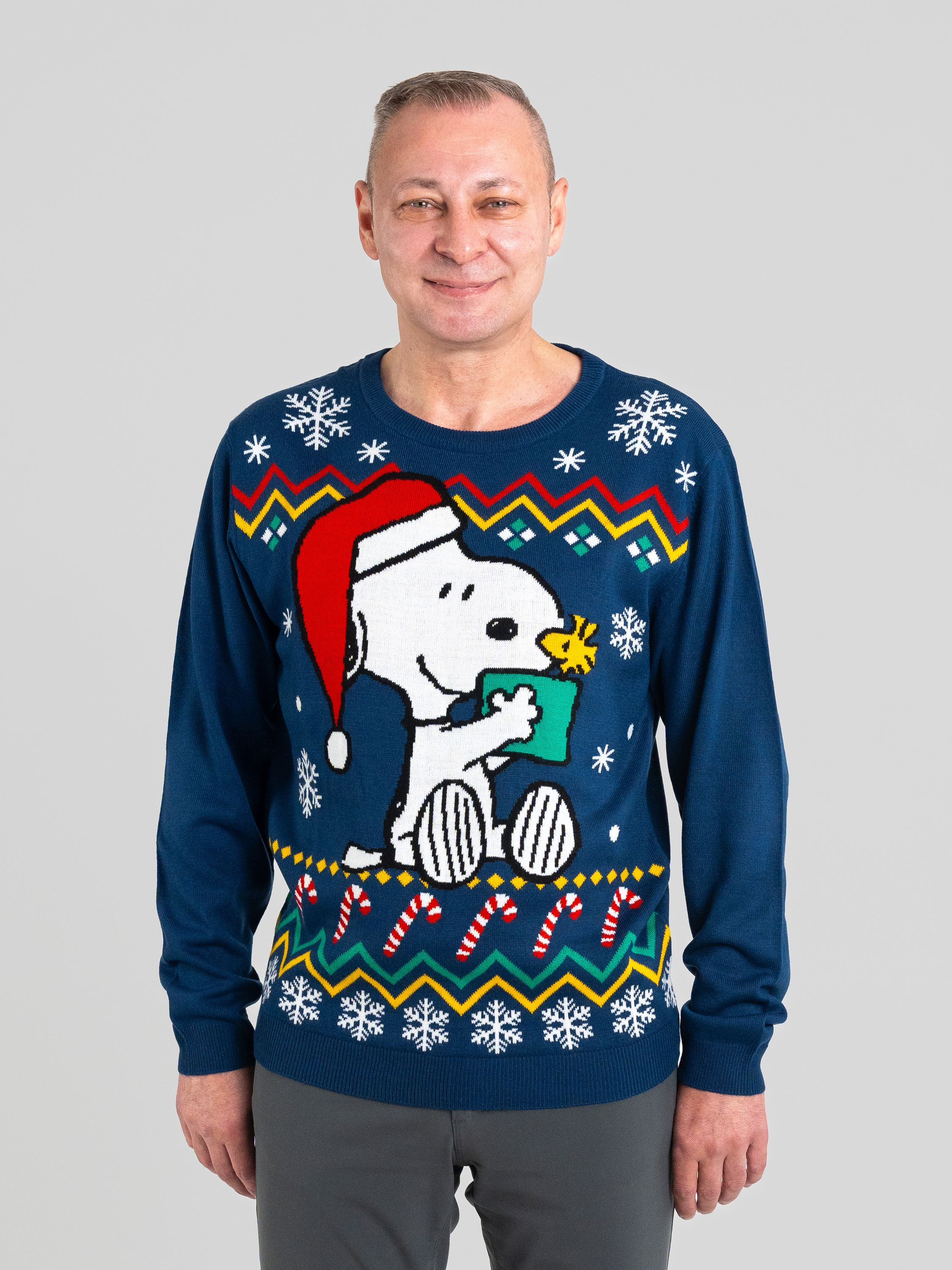 Ein Mann in einem blauen Snoopy-Weihnachtspullover lächelt. Der Pullover zeigt Snoopy und Schneeflocken.