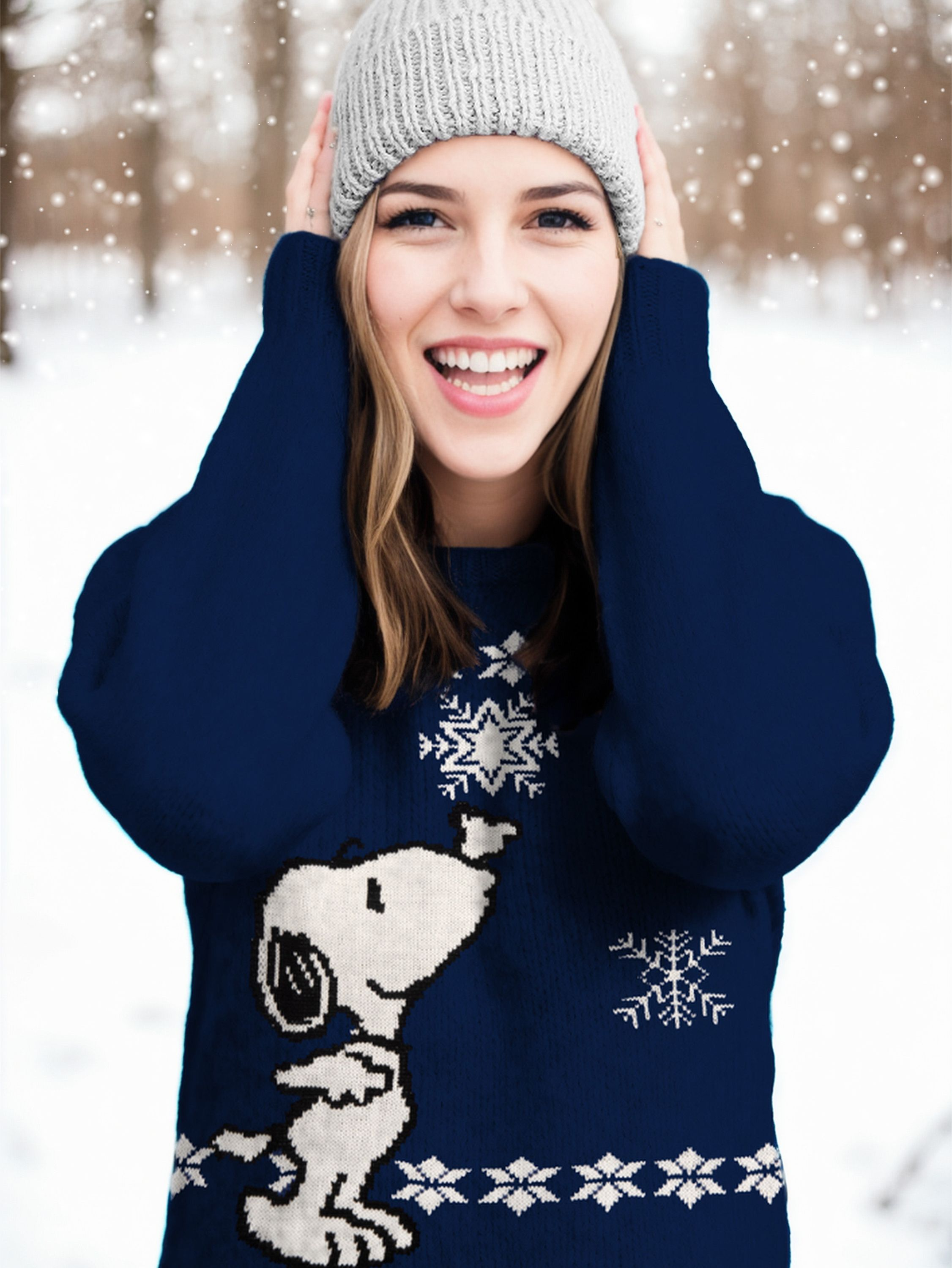 Eine lächelnde Frau in einem marineblauen Pullover mit Snoopy-Design und einer grauen Mütze, Schnee im Hintergrund.