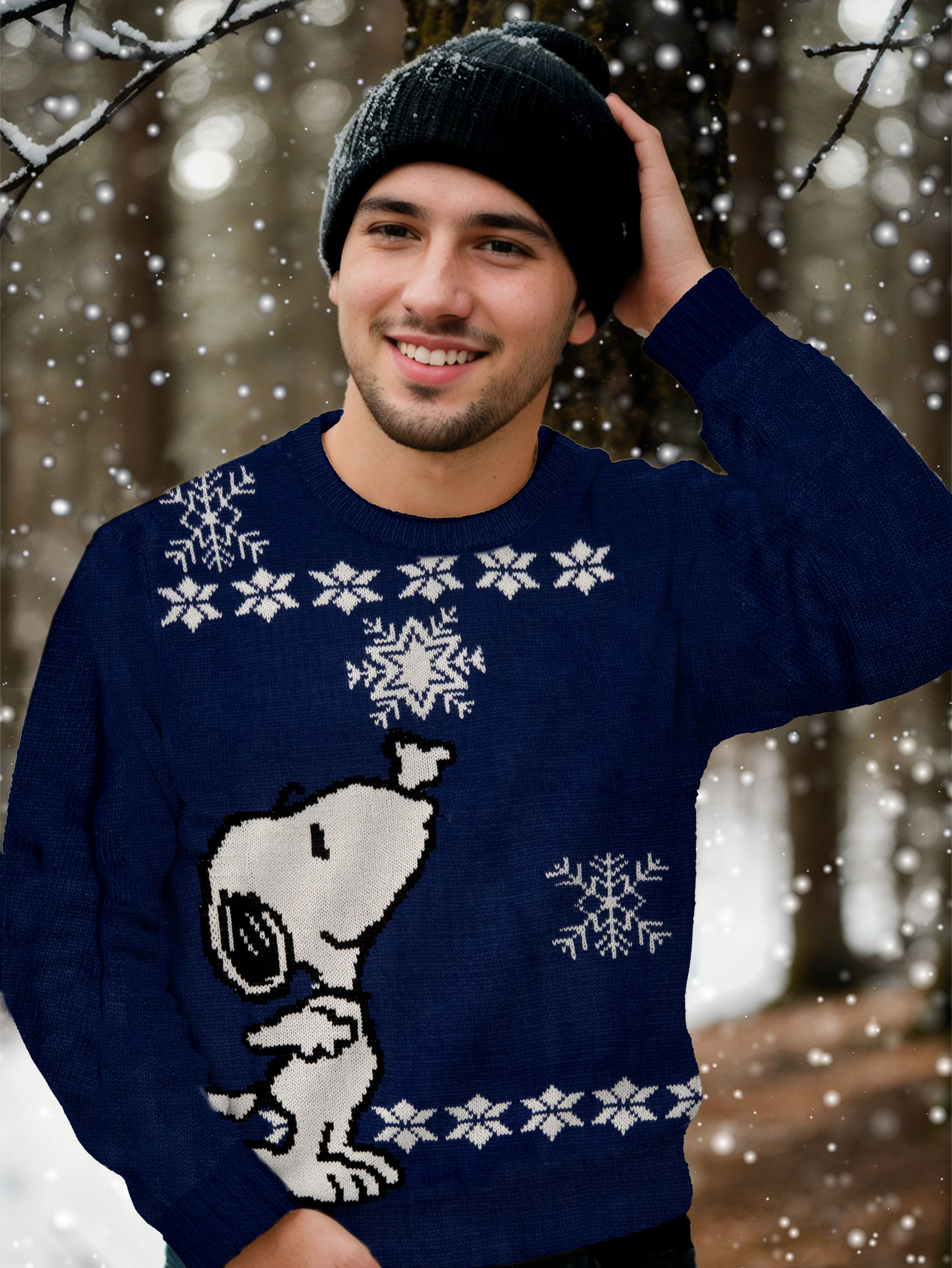 Ein Mann in einem blauen Snoopy-Pullover und einer schwarzen Mütze lächelt in einem verschneiten Wald.
