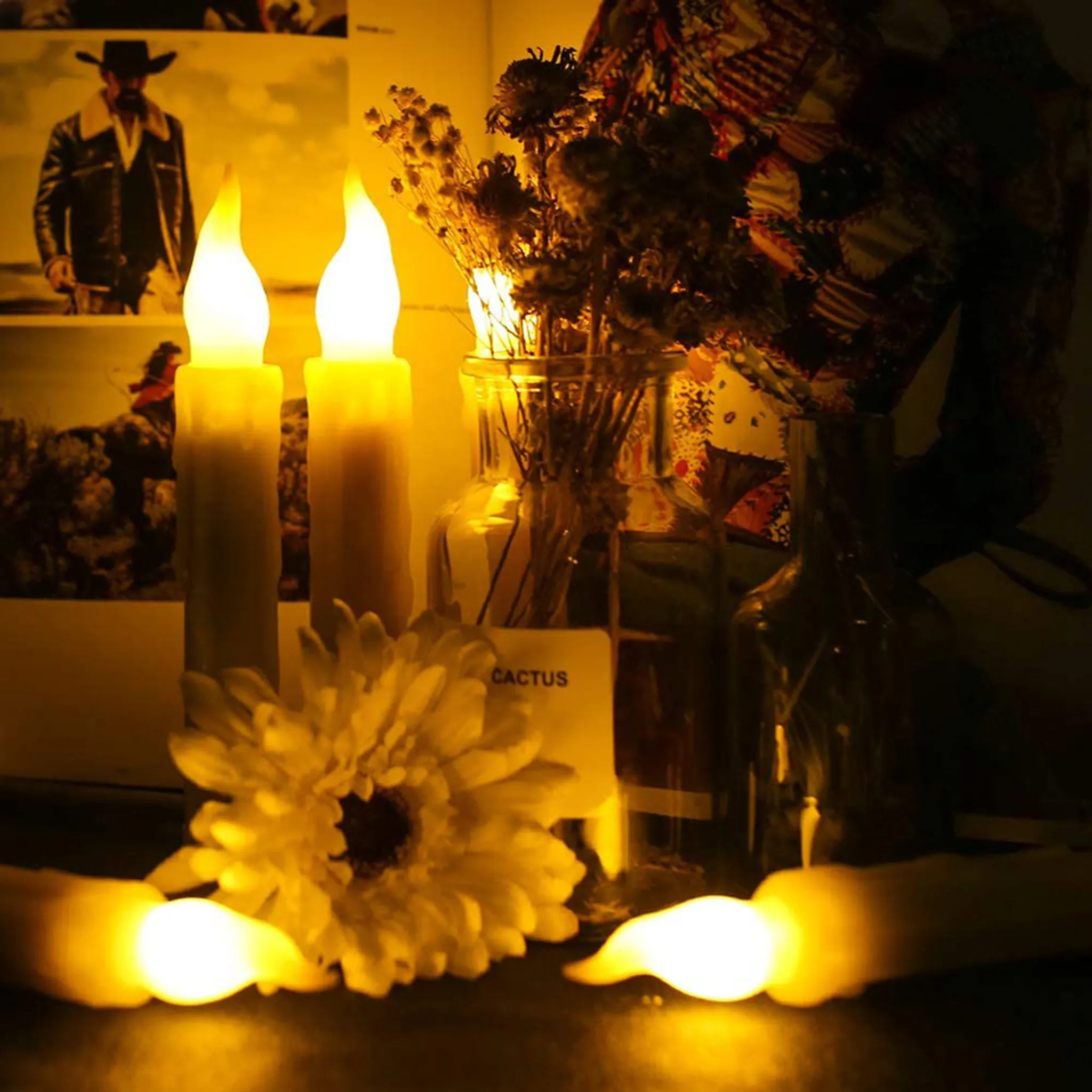Zwei brennende Kerzen, eine Blume, getrocknete Pflanzen in einem Glas und ein Bild eines Mannes mit Cowboyhut sind zu sehen.