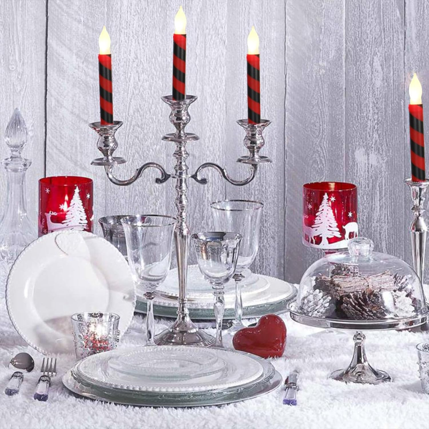 Festliche Tischdekoration mit rot-schwarzen Kerzen, weißen Tellern, Gläsern und einem herzförmigen Ornament auf einer weißen Oberfläche.