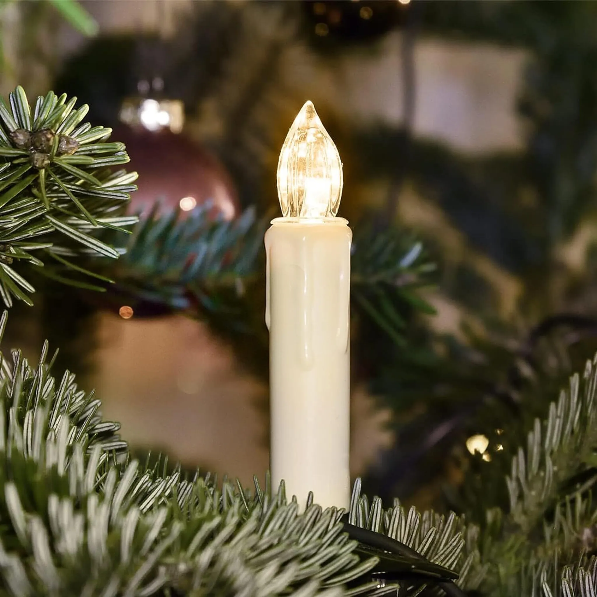 Eine brennende weiße Kerze steht zwischen grünen Tannennadeln. Der Hintergrund ist ein verschwommener Weihnachtsbaum.