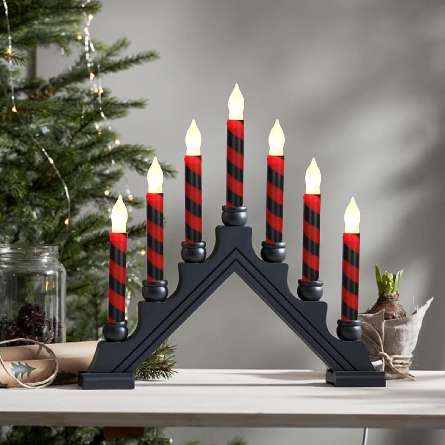 Ein Kerzenhalter mit sieben rot-schwarz gestreiften Kerzen. Ein Weihnachtsbaum und andere Dekorationen sind im Hintergrund.