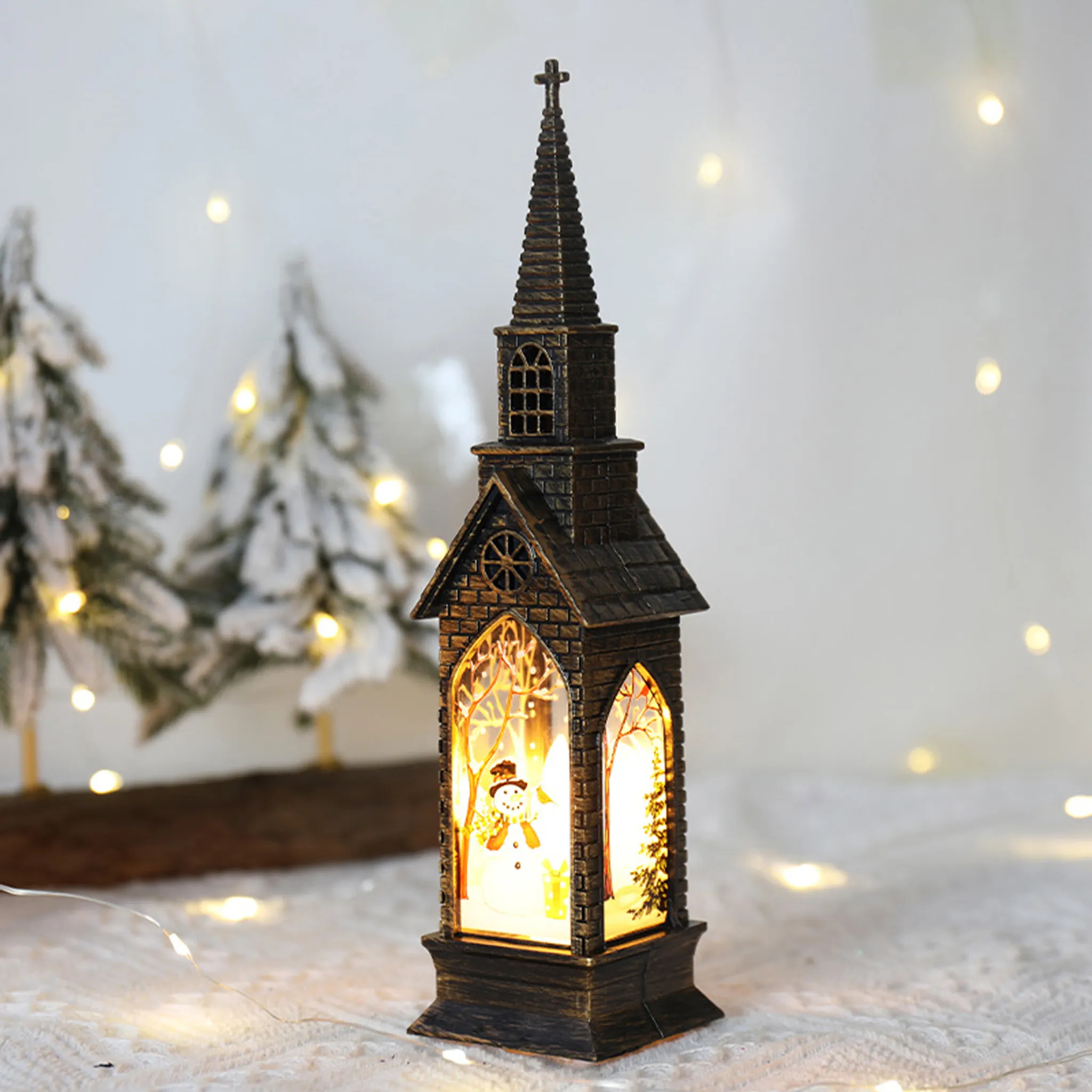 Eine kirchenförmige Weihnachtsdekoration mit einem Schneemann darin. Sie hat ein Kreuz oben und ist beleuchtet. Verschneite Bäume im Hintergrund.