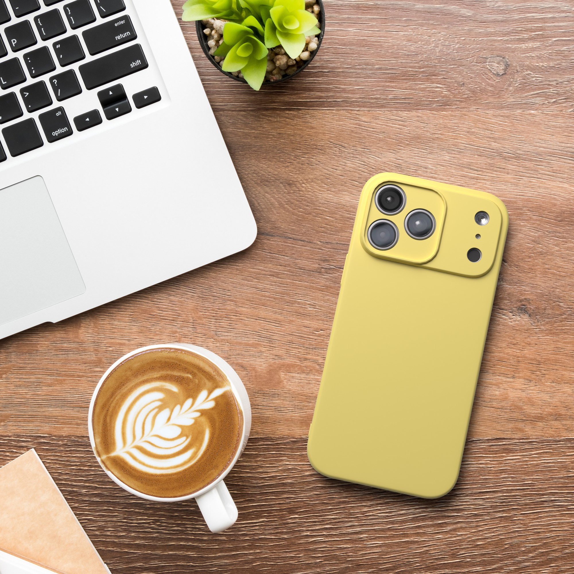 Auf einem Holztisch liegen Laptop, Kaffeetasse mit Latte Art und eine gelbe Handyhülle mit Pflanze.