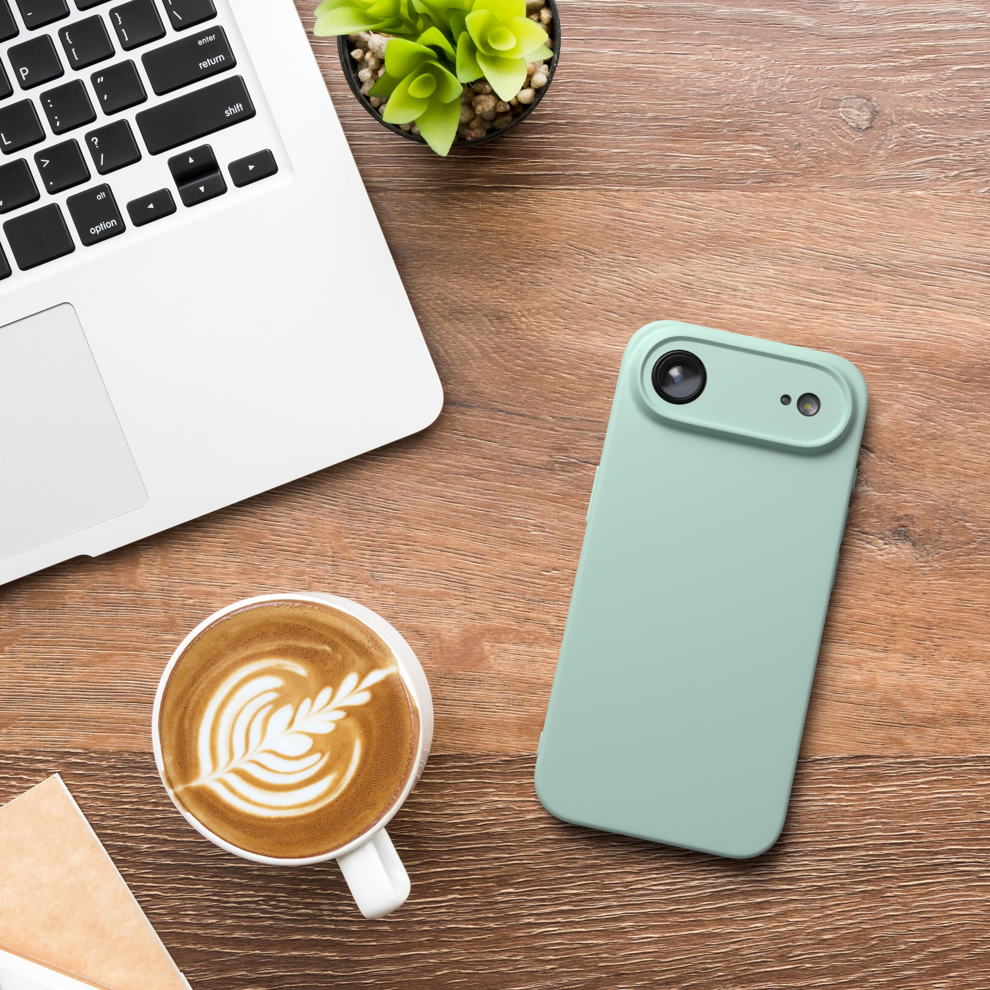 Auf einem Holztisch liegen ein Laptop, ein Telefon, eine Kaffeetasse und eine kleine Pflanze. Der Kaffee hat Latte Art.