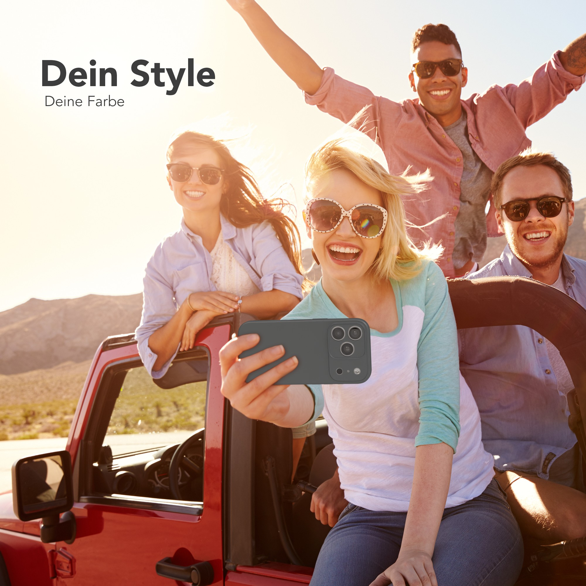 Freundesgruppe in einem roten Jeep macht ein Selfie. Einer hält eine Handyhülle. Helles Sonnenlicht und Wüstenhintergrund.