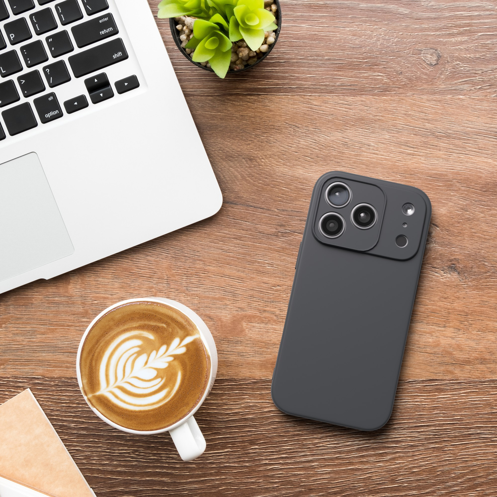 Ein Laptop, ein Telefon, ein Kaffee und eine Pflanze auf einem Holzschreibtisch. Der Kaffee hat Latte Art. Das Telefon ist dunkelgrau.