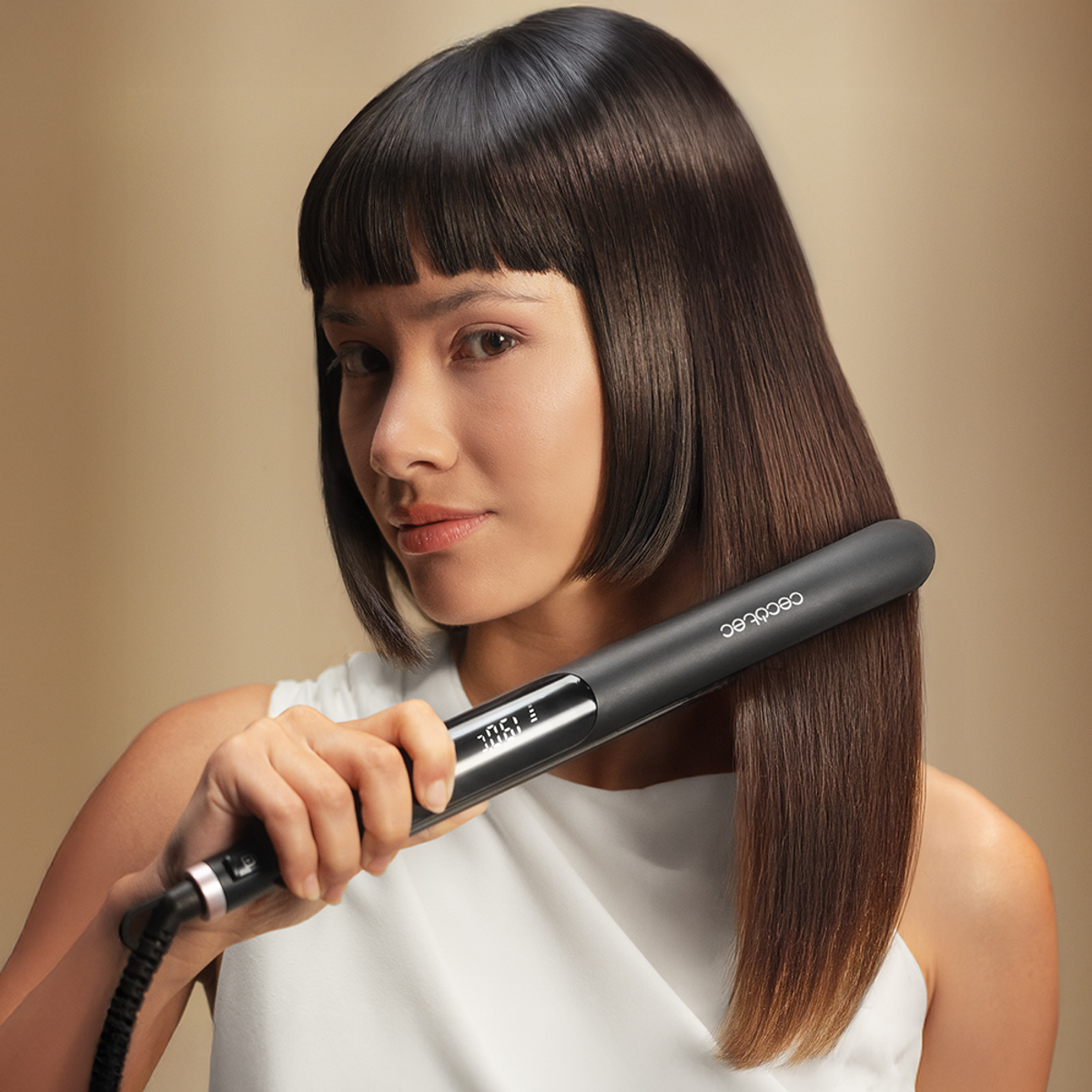 Una donna usa una piastra nera sui suoi capelli scuri e lisci. Indossa una maglietta bianca e guarda la fotocamera.