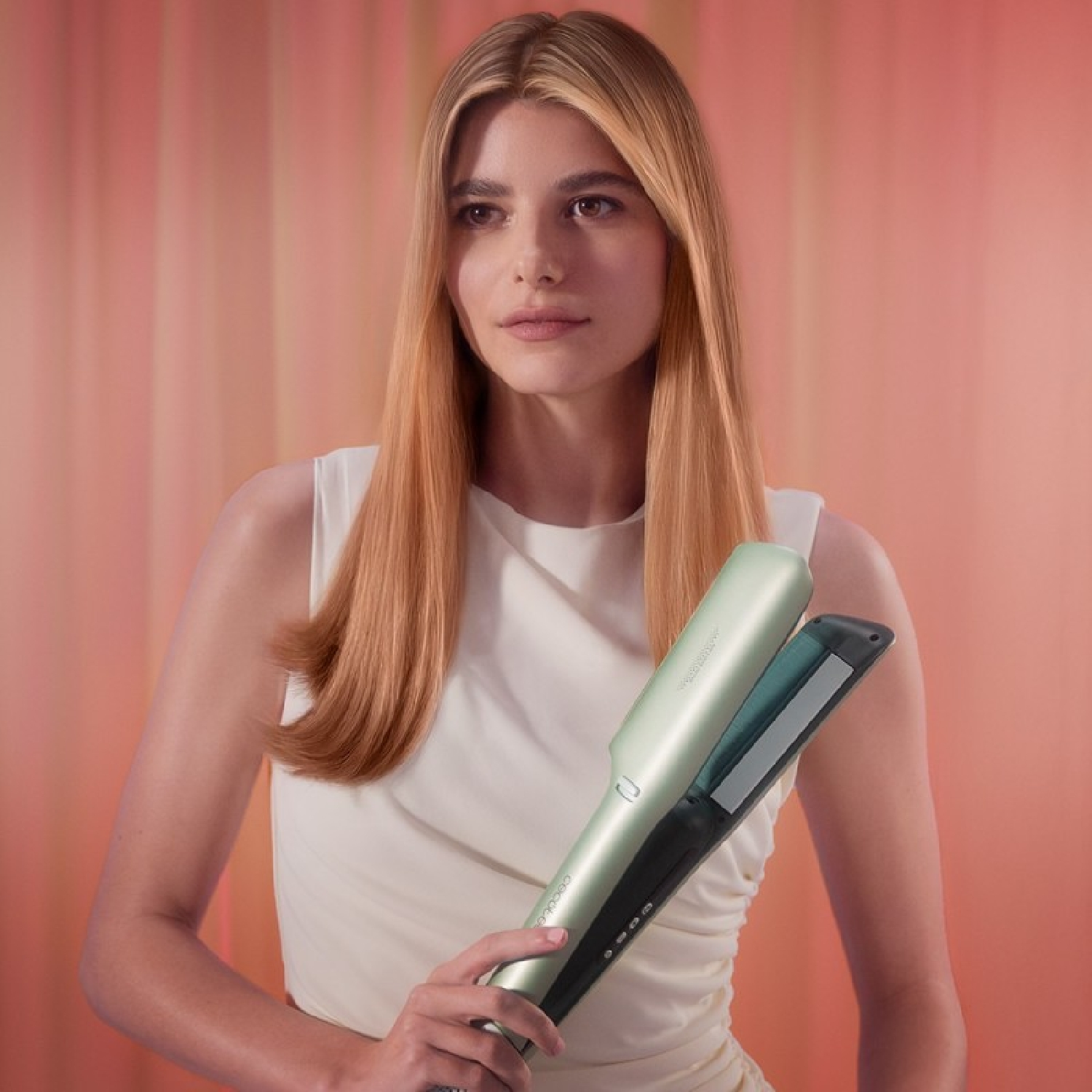 Una mujer sostiene una plancha de pelo verde. Tiene el pelo largo y rubio y viste un vestido blanco. El fondo es rosa.