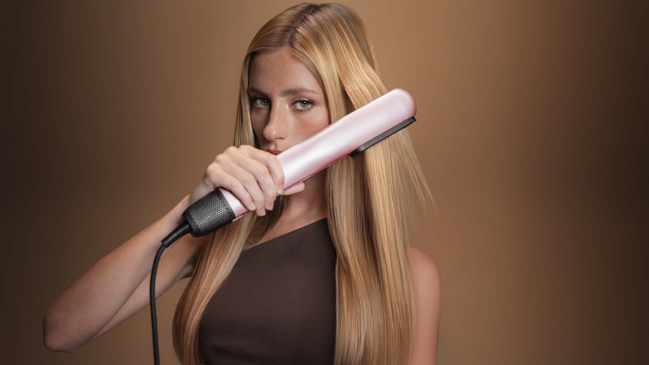 Een vrouw met blond haar steilt haar haar met een roze en zwarte stijltang. Ze draagt een bruine top en kijkt naar de camera.
