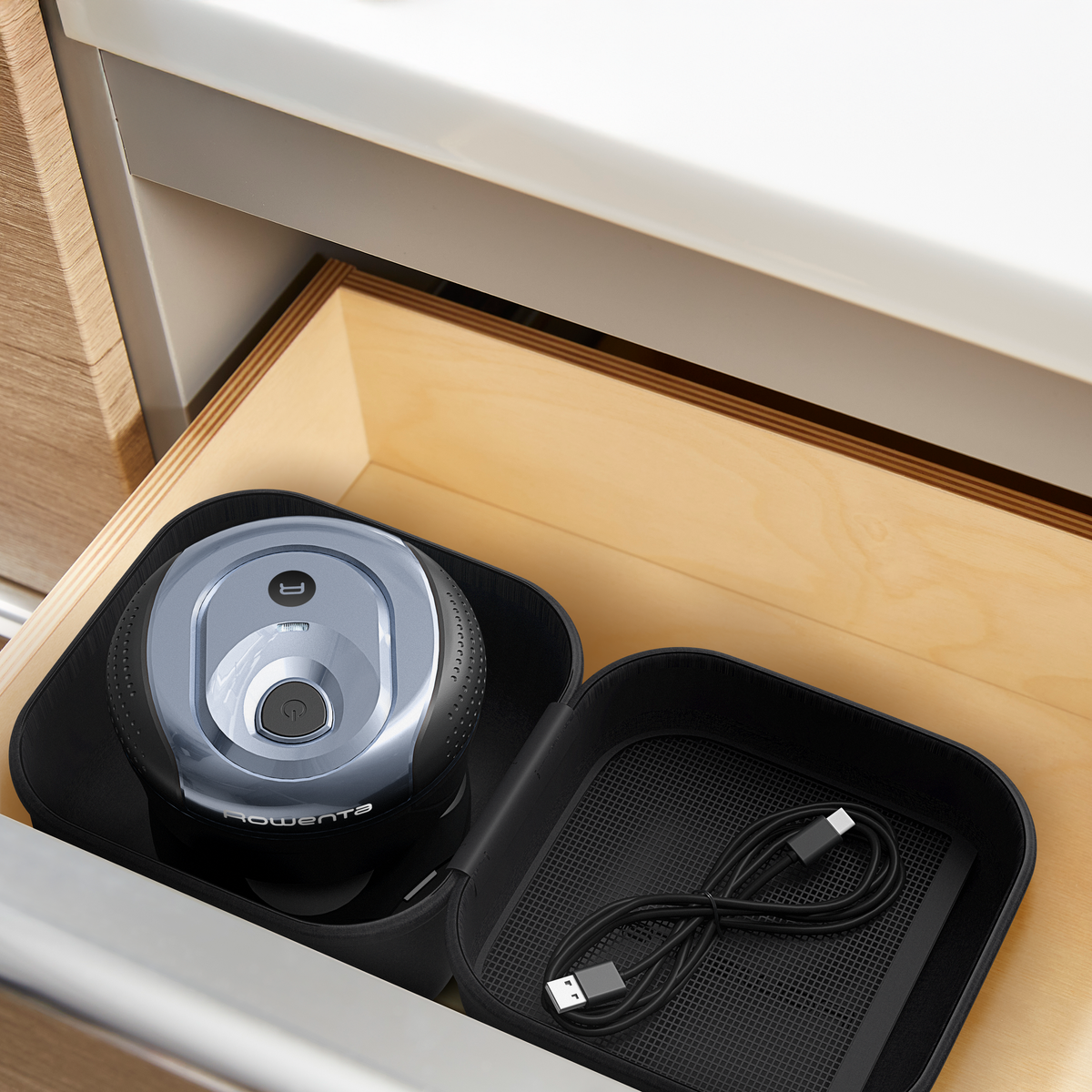 Un piccolo aspirapolvere Rowenta argentato e nero si trova in una custodia nera aperta all'interno di un cassetto di legno. Un cavo USB è anche nella custodia.