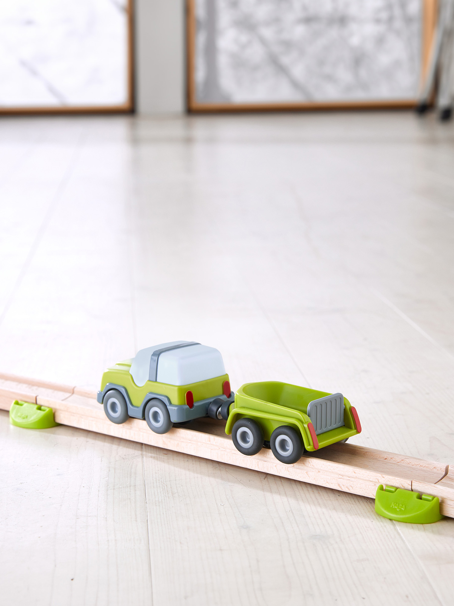 Zielony samochodzik z przyczepą stoi na drewnianym torze. Tło przedstawia rozmyte obrazy i drewnianą podłogę.