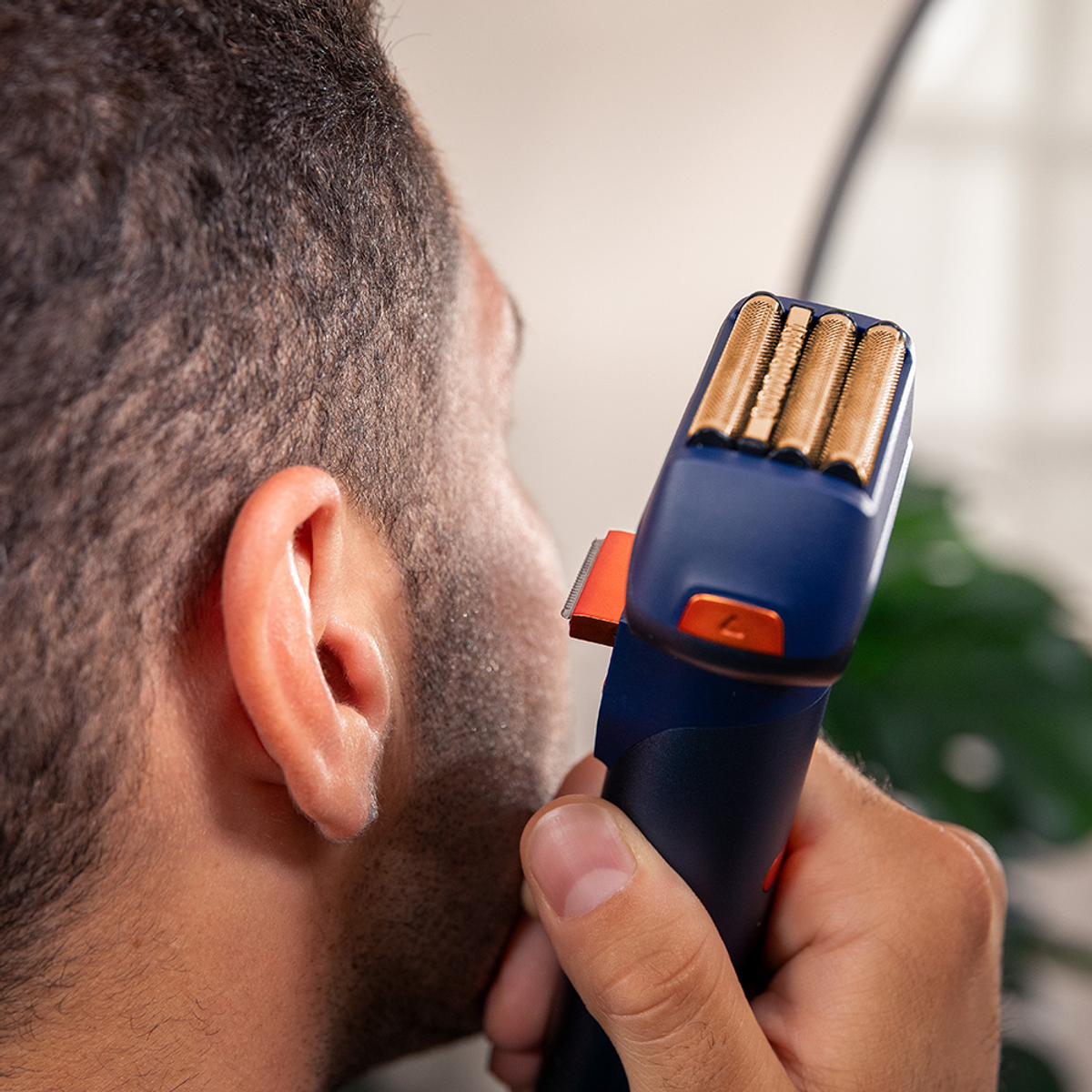 Un uomo si rade la barba con un rasoio elettrico. Il rasoio è blu e arancione. Il viso e l'orecchio dell'uomo sono visibili.
