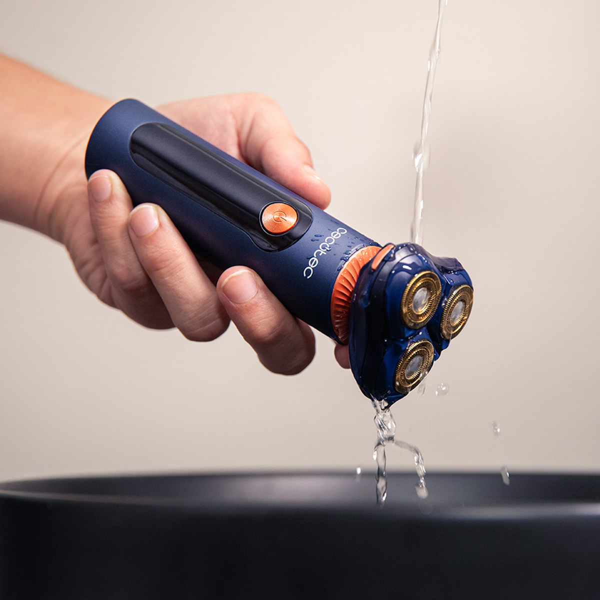 Una mano tiene un rasoio elettrico blu e arancione sotto l'acqua corrente. Il rasoio ha tre testine rotanti dorate.
