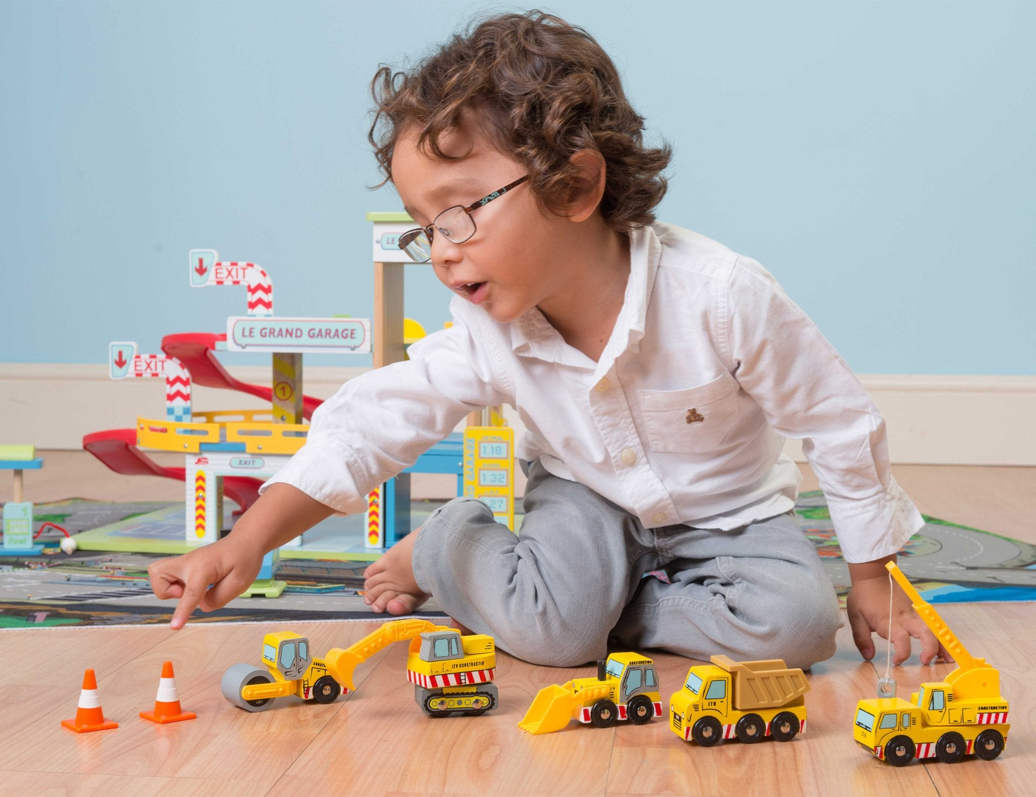Un niño con gafas juega con vehículos de construcción de juguete. Un garaje de juguete está en el fondo.