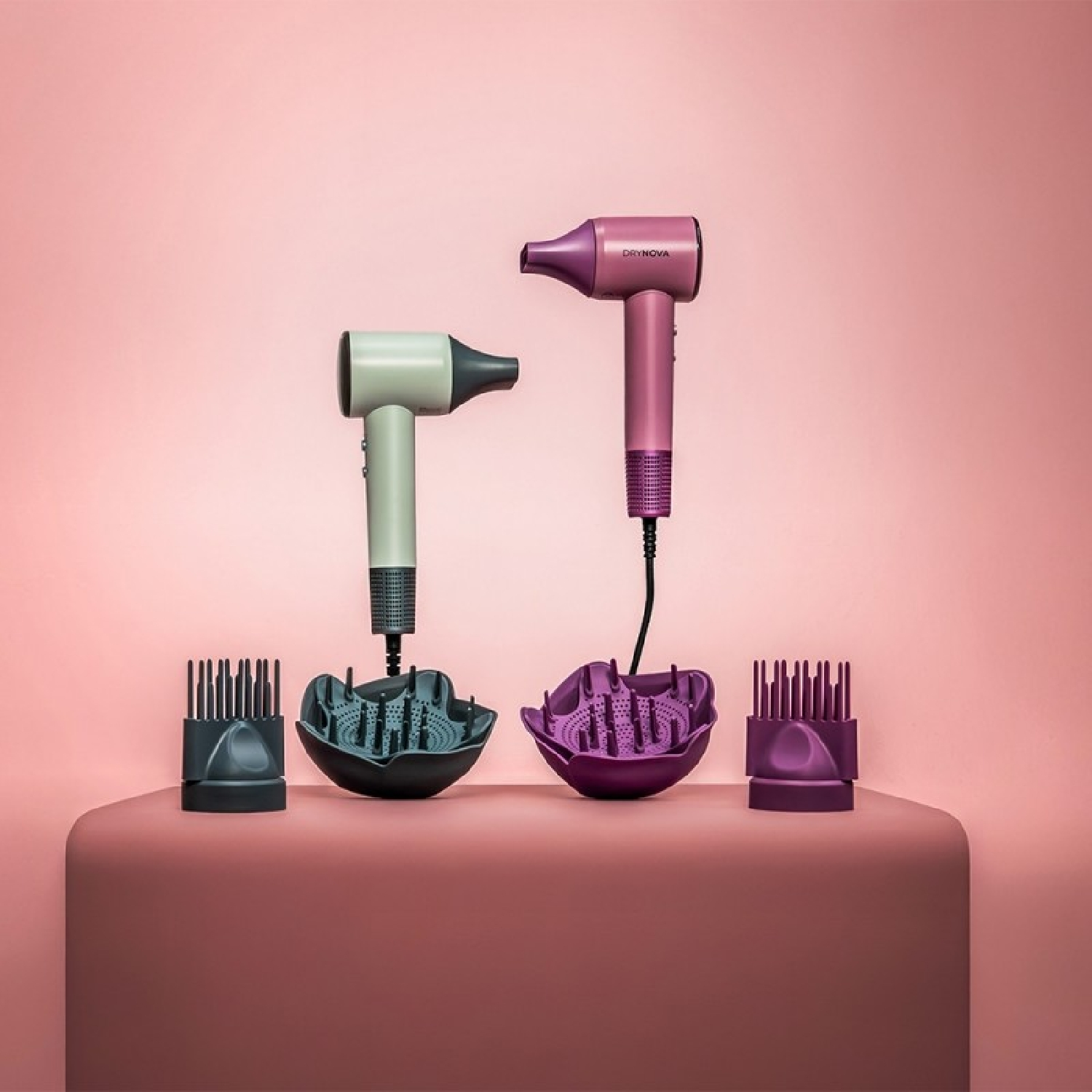 Zwei Haartrockner, Aufsätze und kammartige Accessoires auf einer rosa Oberfläche. Hintergrund ist ein rosa Farbverlauf. Ein Haartrockner ist beige.
