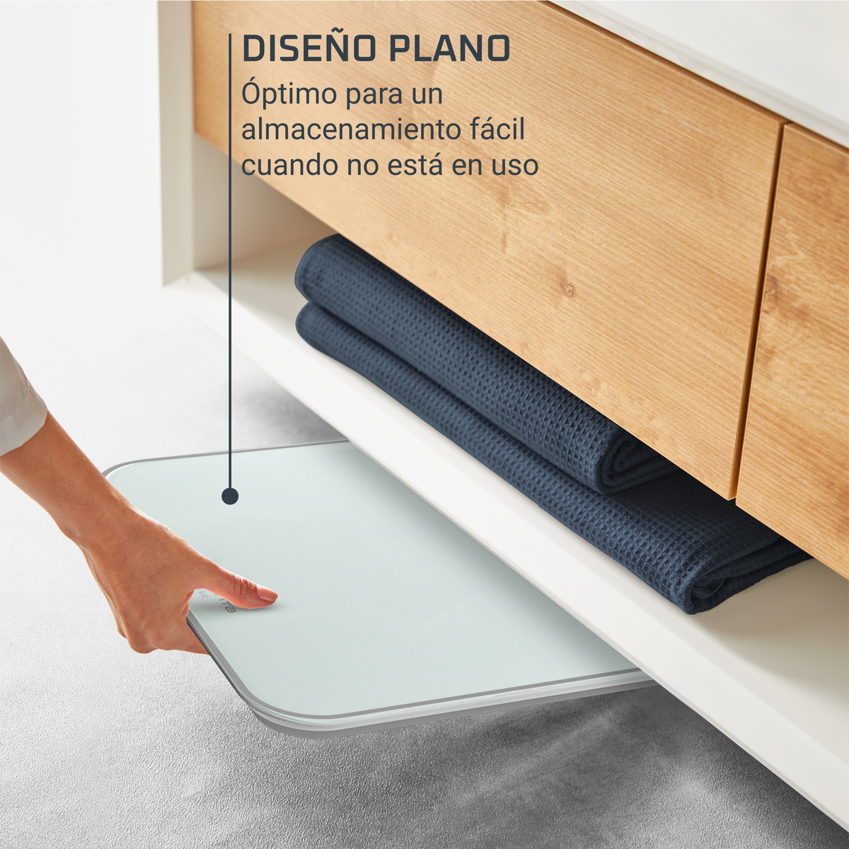 Eine Hand zieht eine weiße Waage unter einem Holzschrank hervor. Zwei blaue Handtücher sind auf dem Regal gefaltet. Text auf Spanisch ist sichtbar.