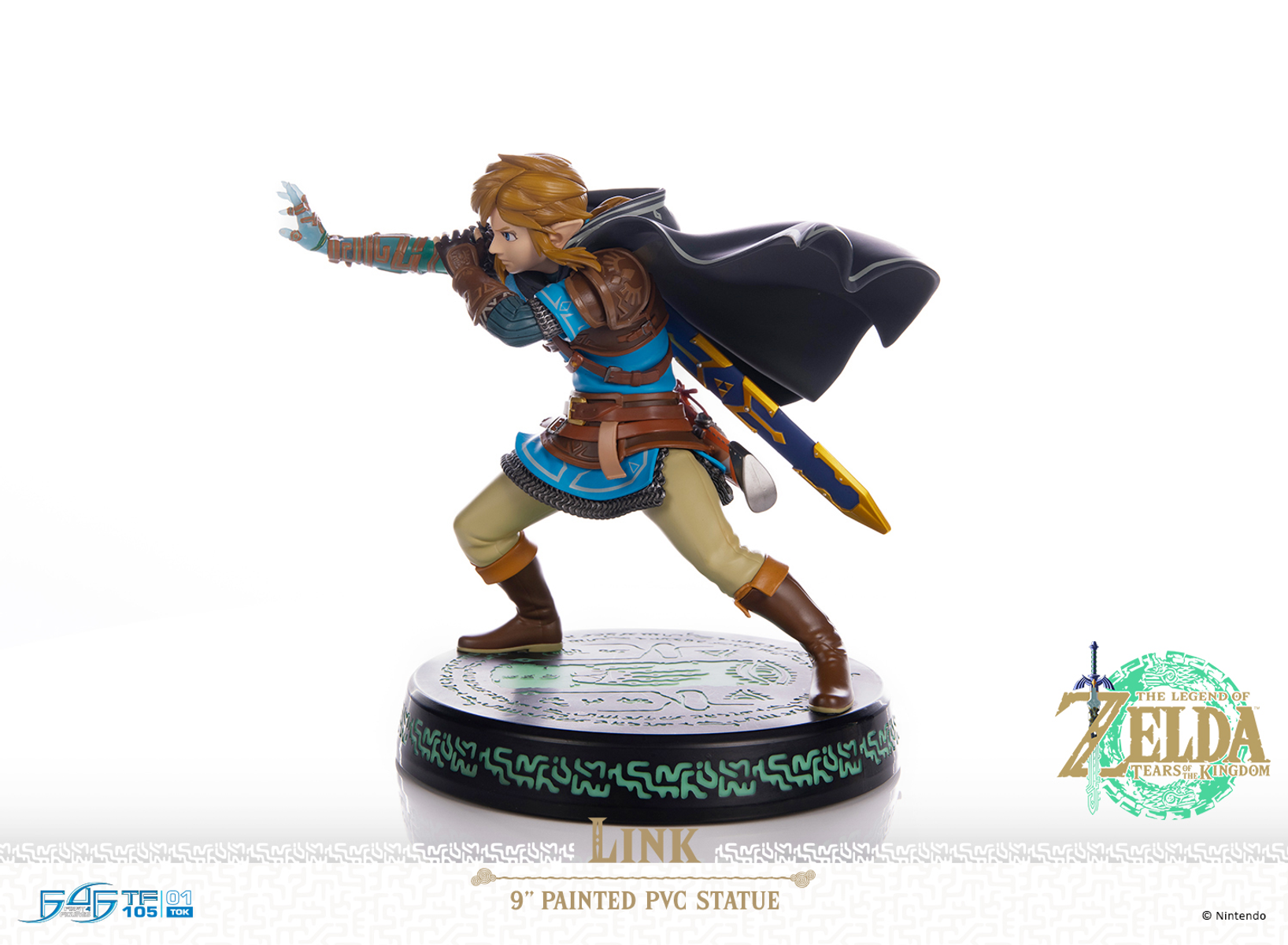 Estatua de Link en pose de acción. Viste armadura azul y marrón, una capa negra y empuña una espada. La base es negra con diseños verdes.