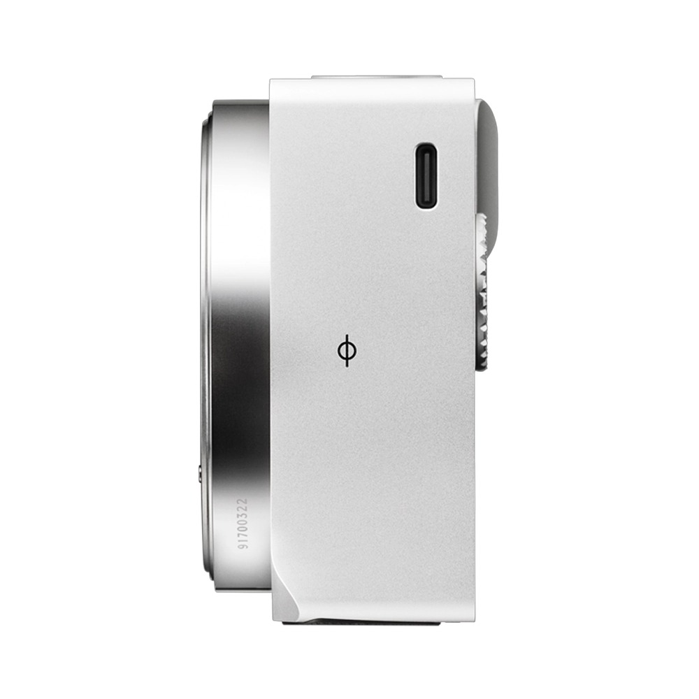 Una fotocamera digitale argentata è mostrata di profilo. Il corpo macchina è rettangolare con un obiettivo e diversi pulsanti. Lo sfondo è bianco.