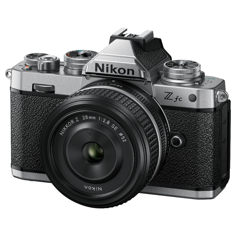 Een zilveren en zwarte Nikon Z fc camera met een lens bevestigd. De lens zegt Nikkor Z 28mm 1:2.8 SE. De camera staat op een wit oppervlak.