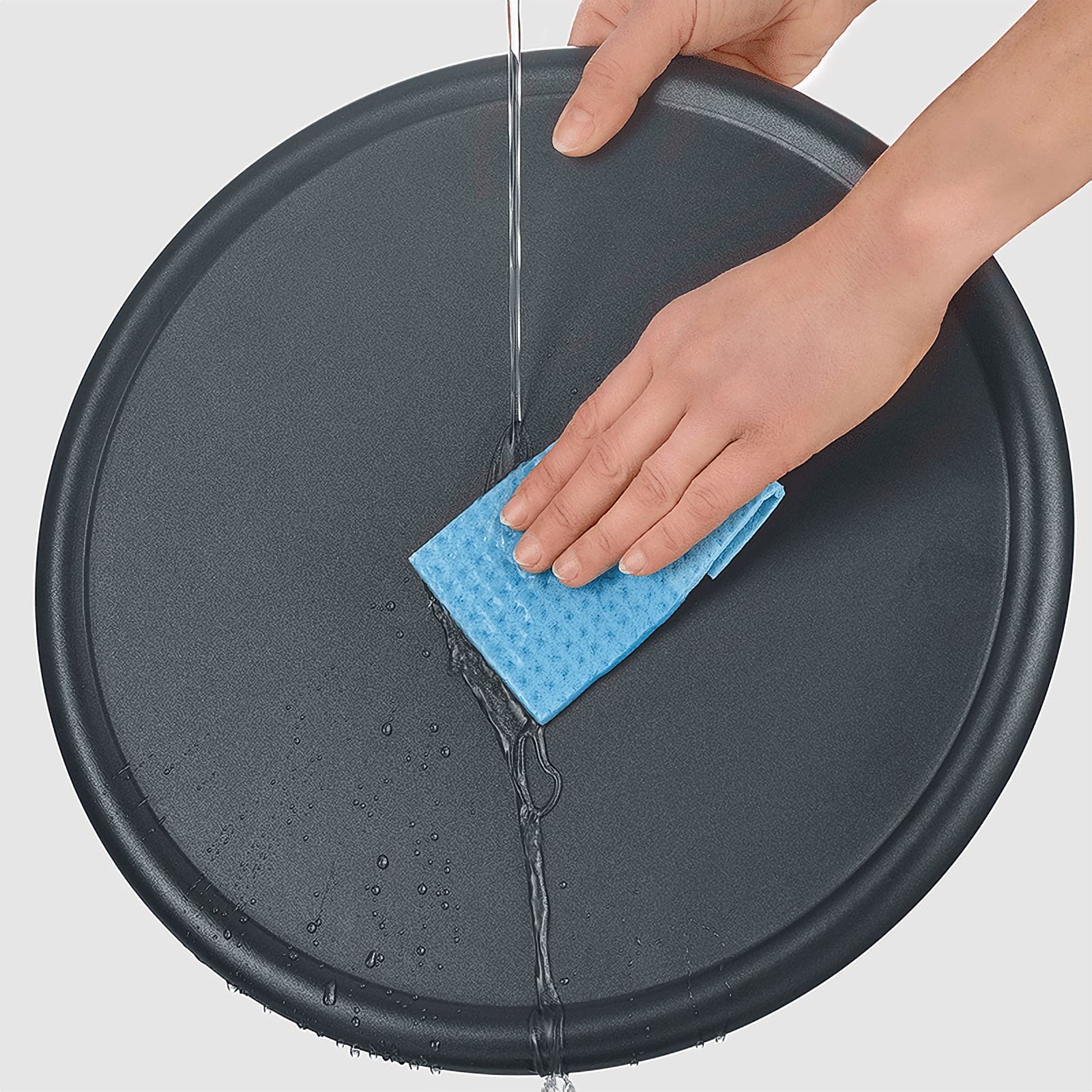 Osoba myje ciemnoszarą tacę niebieską gąbką pod bieżącą wodą. Widoczne krople wody.