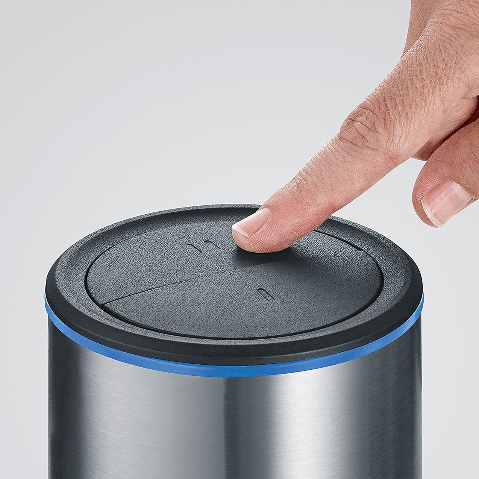 Un dito preme un pulsante sul coperchio di un cestino. Il cestino è argentato con un anello blu e un coperchio nero.