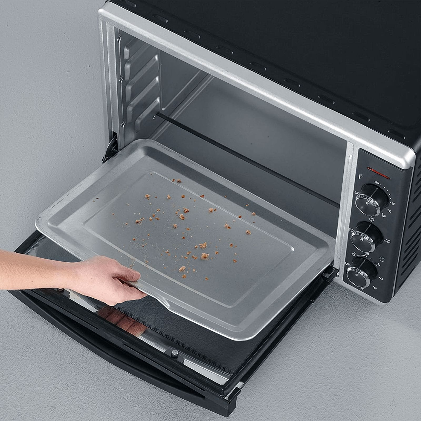 Un horno con la puerta abierta. Una mano sostiene una bandeja con migas. El horno es negro y plateado.